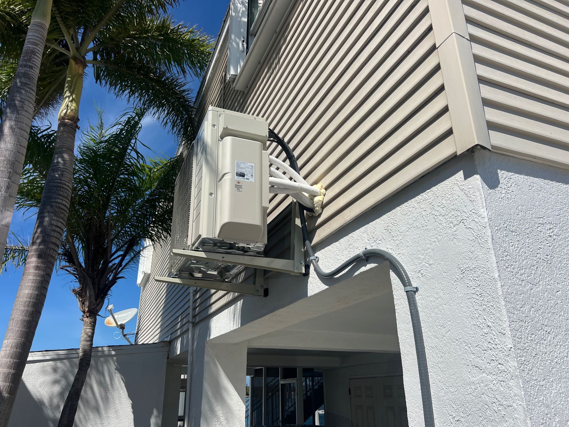 Exterior air conditioning unit mounted on a building's wall, with electrical conduit and a small palm tree visible.