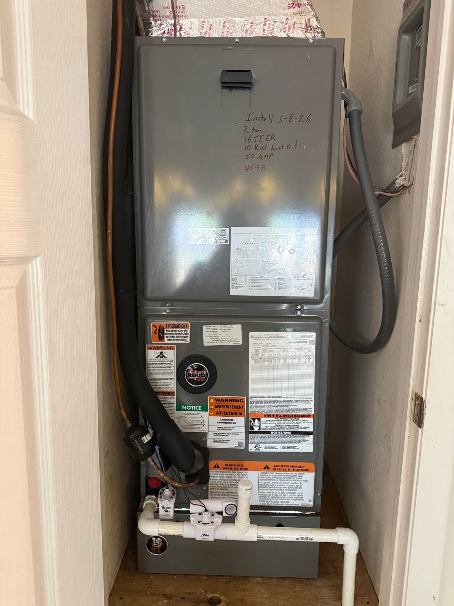 Gray HVAC furnace in a narrow closet with exposed pipes and ducts.