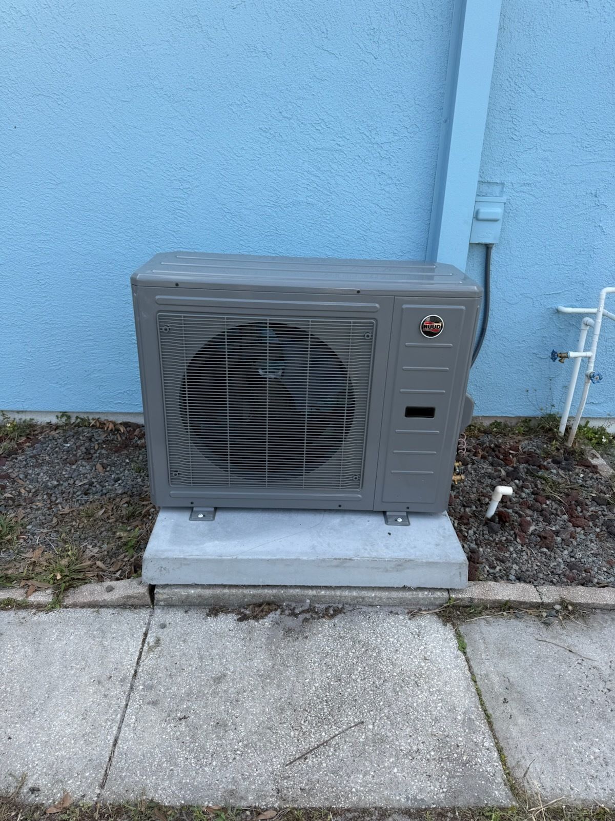 Outdoor gray HVAC unit on a concrete pad against a blue exterior wall beside a sidewalk