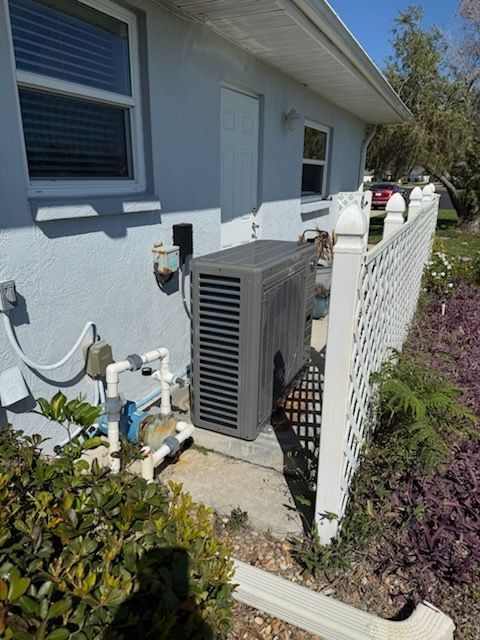 Outdoor HVAC unit beside a house wall and white fence in a sunny garden area