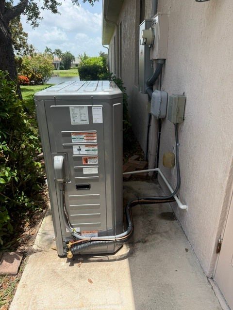 Outdoor HVAC unit beside a house wall with electrical conduits and utility boxes, next to a sidewalk.