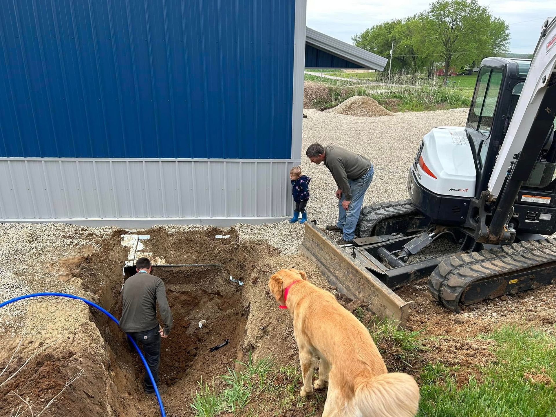 Workers and a dog beside an excavator at a foundation dig next to a blue building