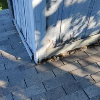 Corner of a weathered wooden structure on a shingled roof, showing damage and debris.