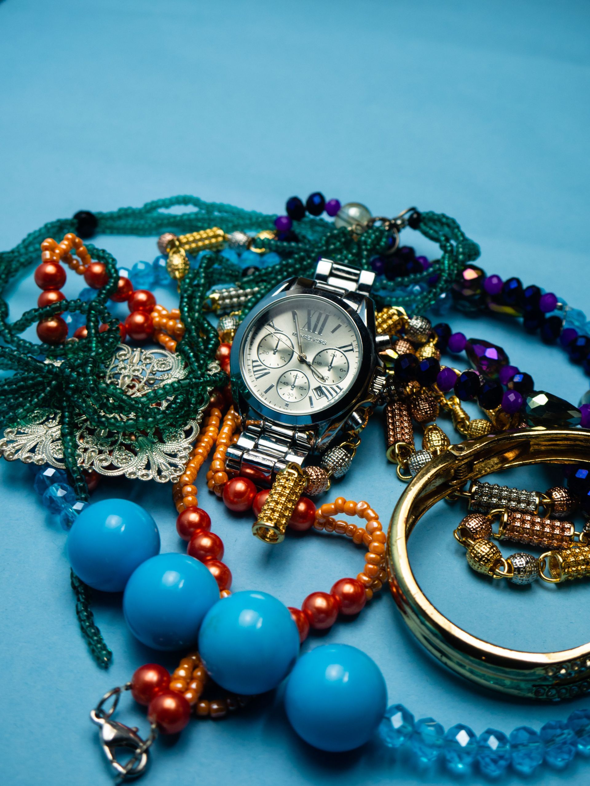 Assorted colorful beaded necklaces and bracelets surrounding a silver wristwatch on a light blue surface