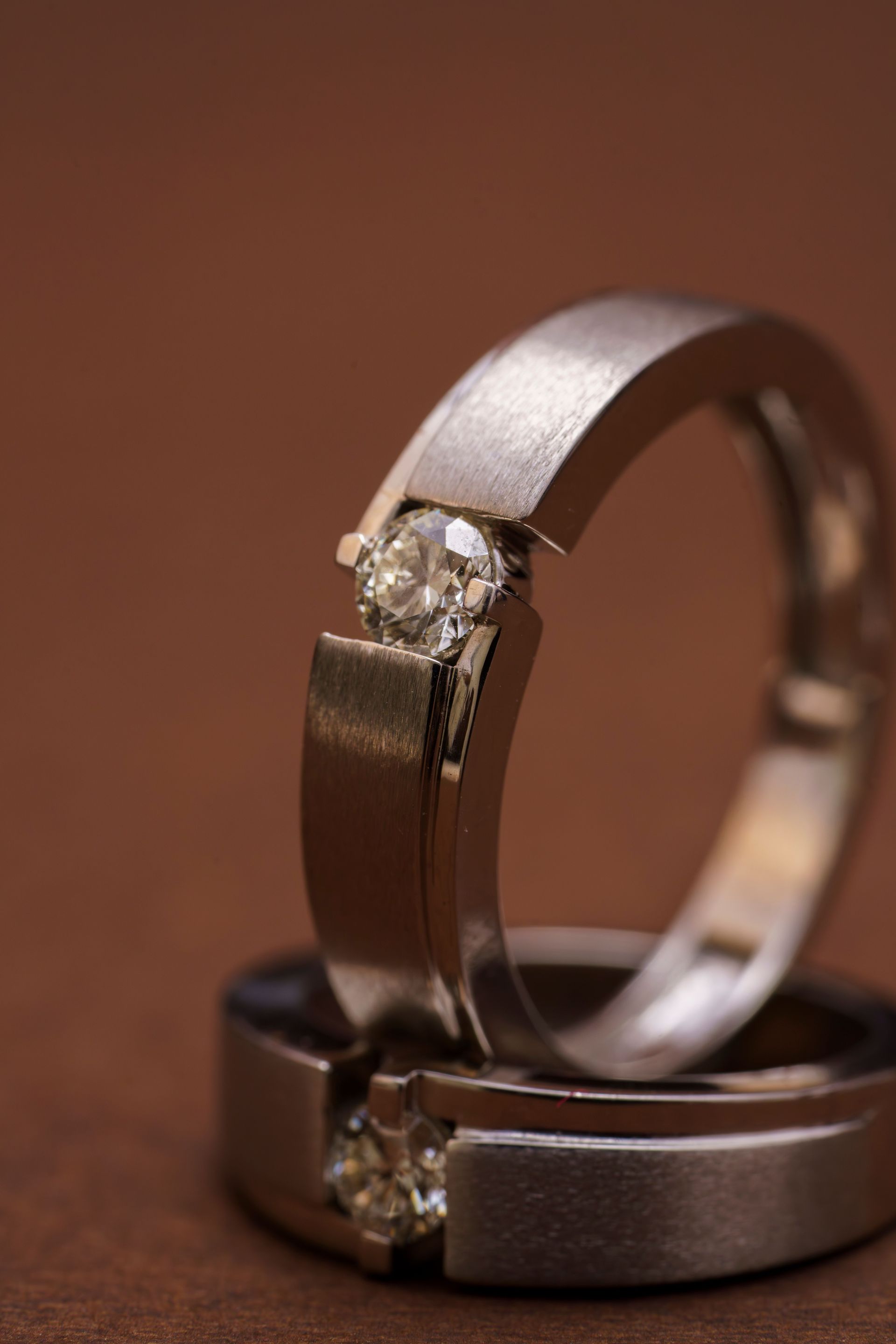 Two silver rings with diamonds, one standing upright and one lying flat, on a brown background