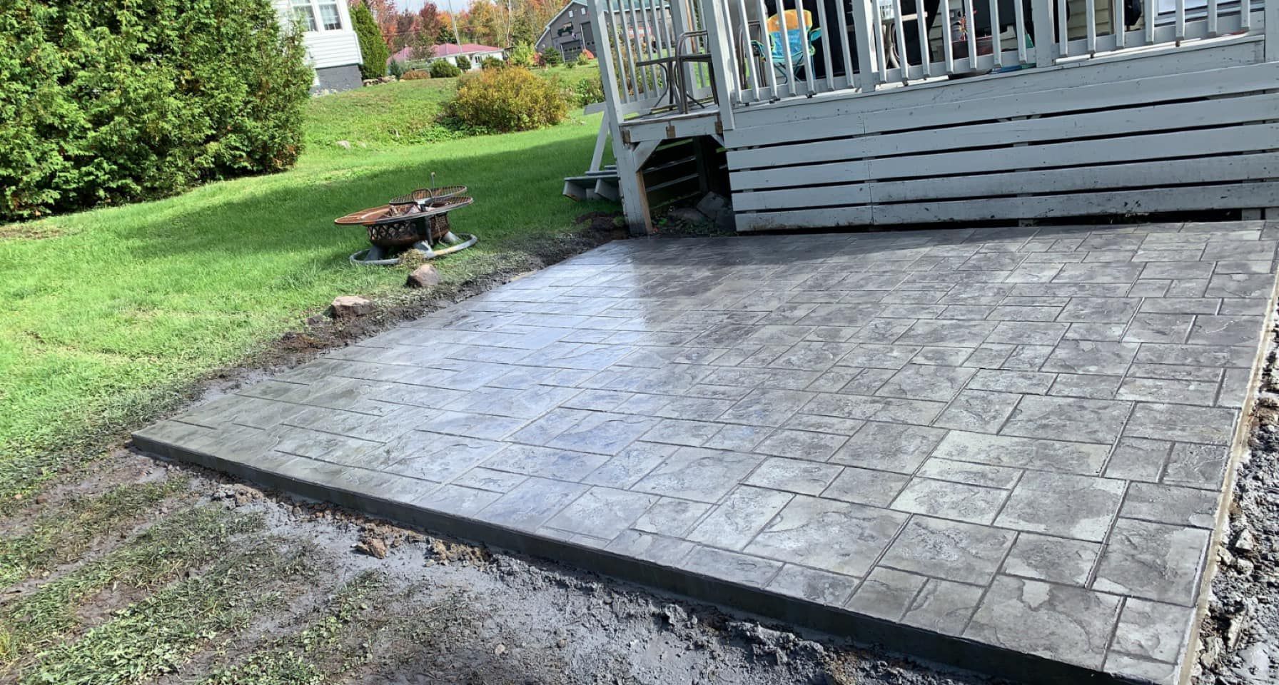 Stamped concrete patio beside a gray deck, bordered by grass and gravel in a backyard.