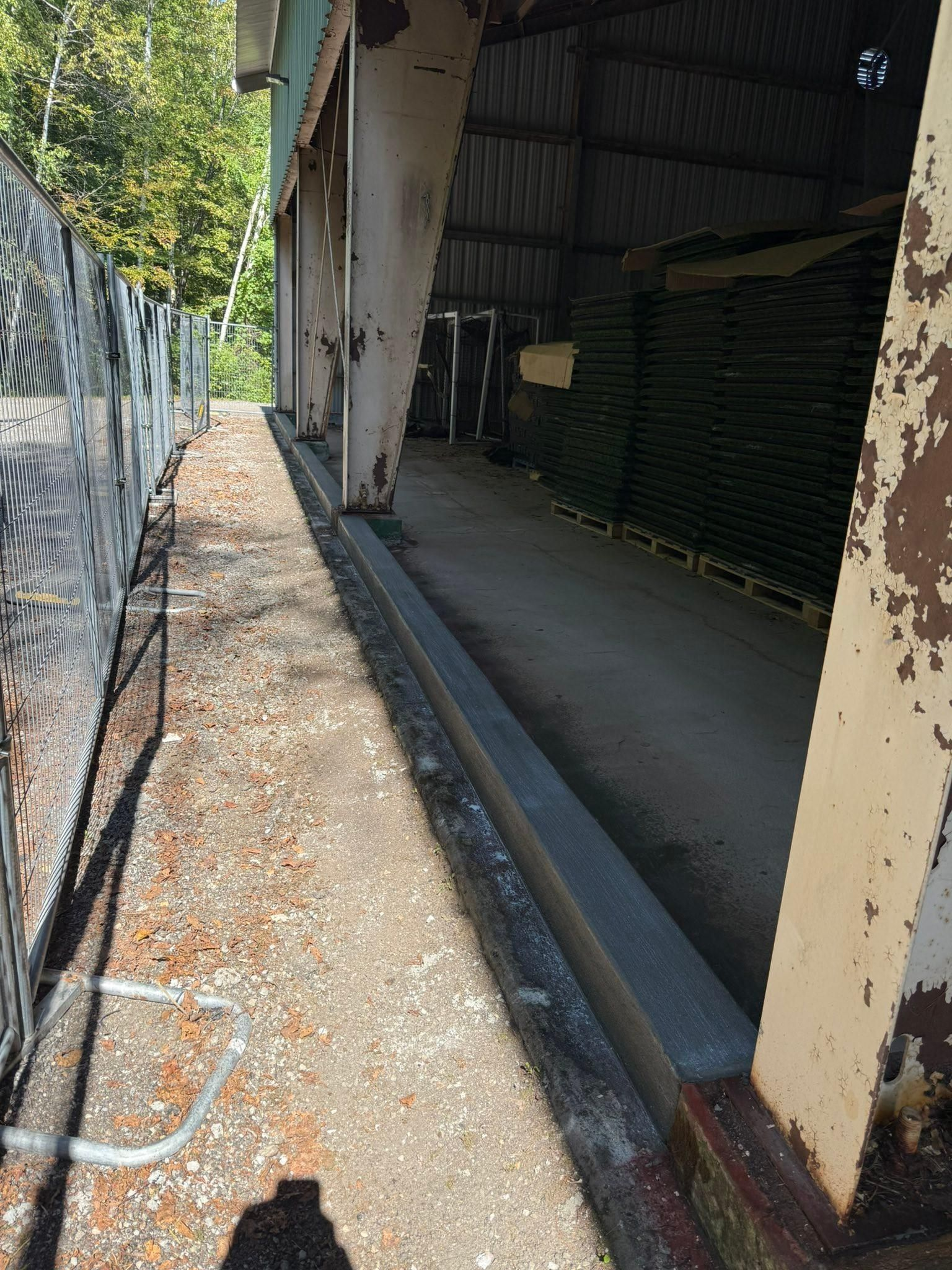 Narrow outdoor walkway beside a warehouse, with chain-link fence on the left and stacked materials inside the open bay.