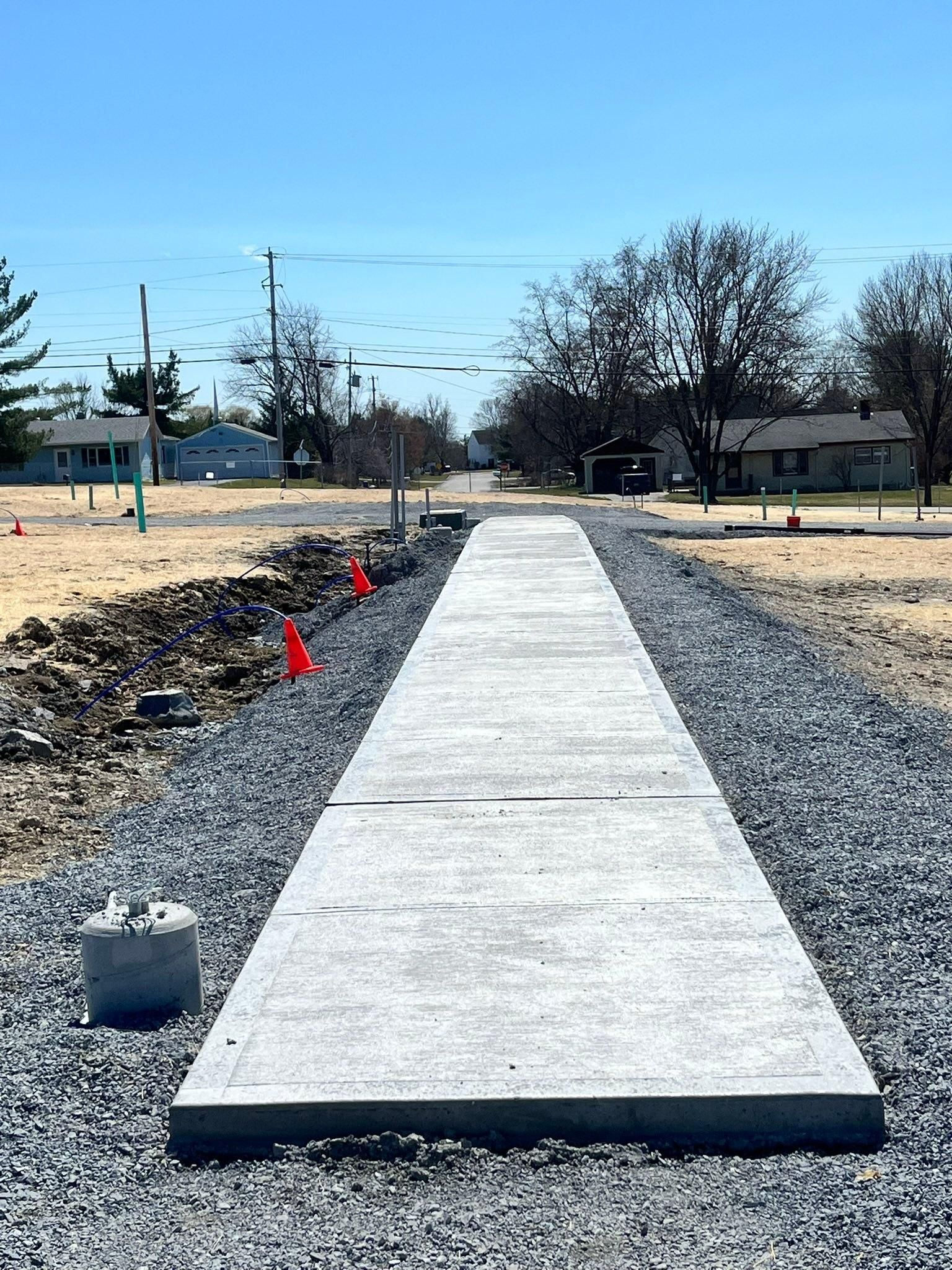 Concrete sidewalk path under construction, with gravel, dirt, and orange markers on both sides.