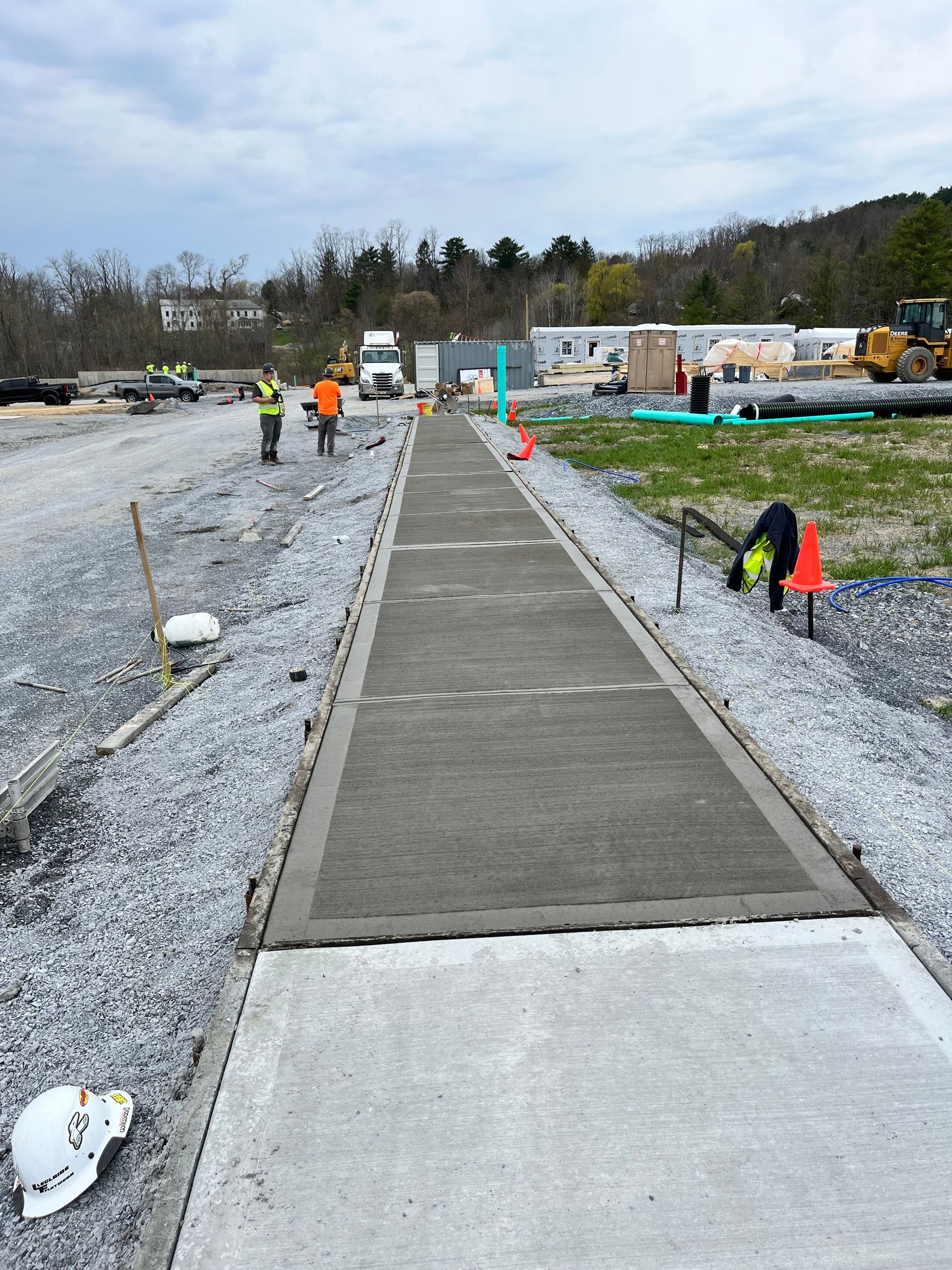 Concrete walkway under construction beside gravel with workers and cones in a cloudy outdoor site.