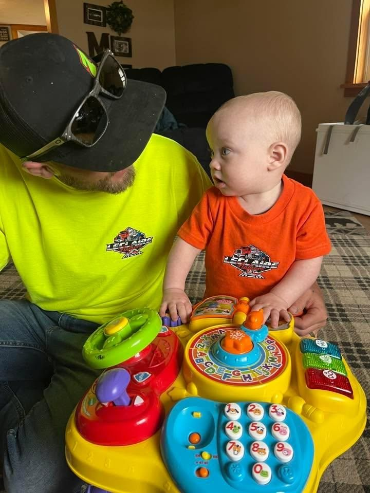 Adult and baby sitting together at a colorful toy table, looking at each other indoors.