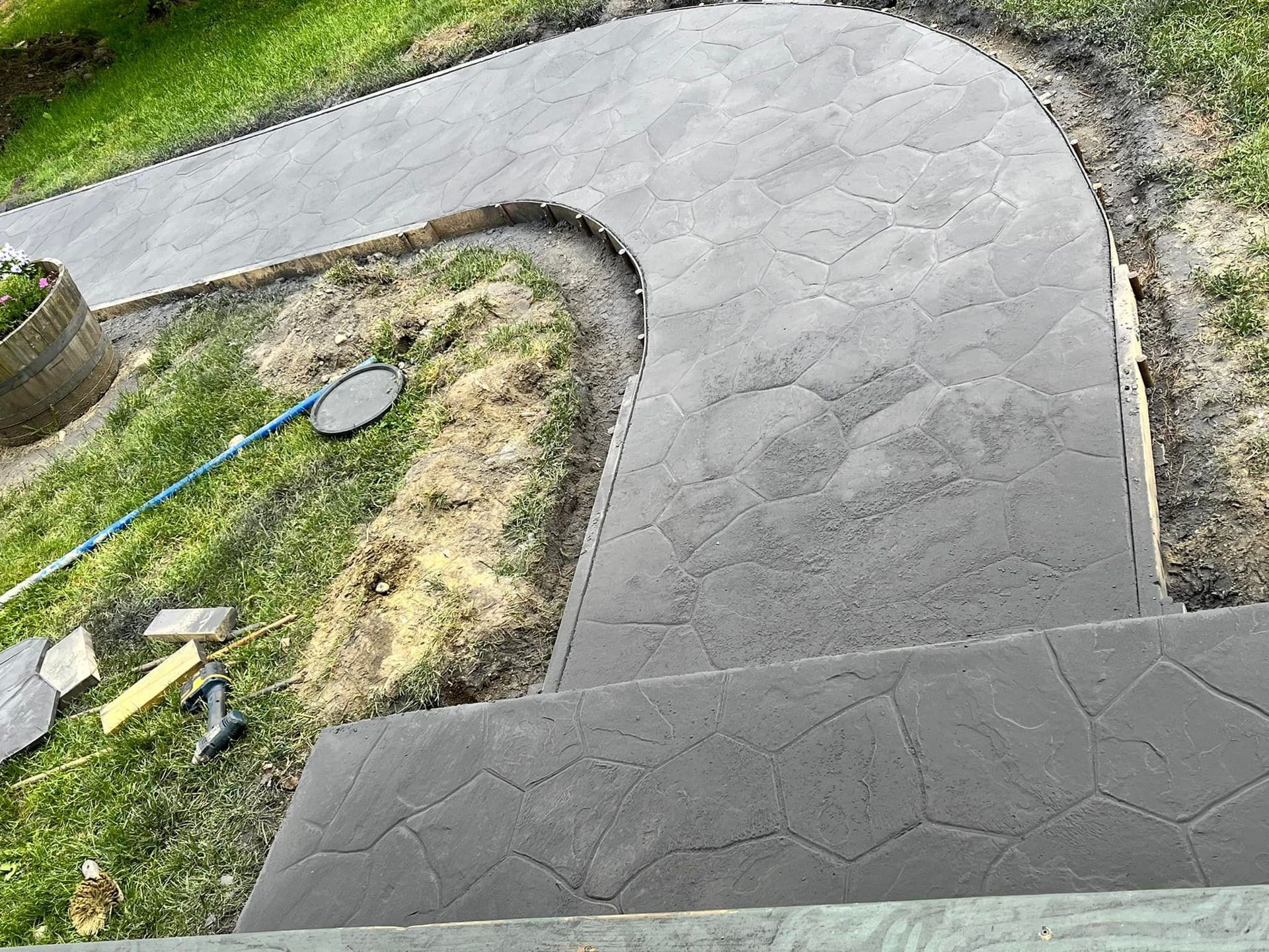 Curved gray concrete walkway beside a grassy yard and garden tools