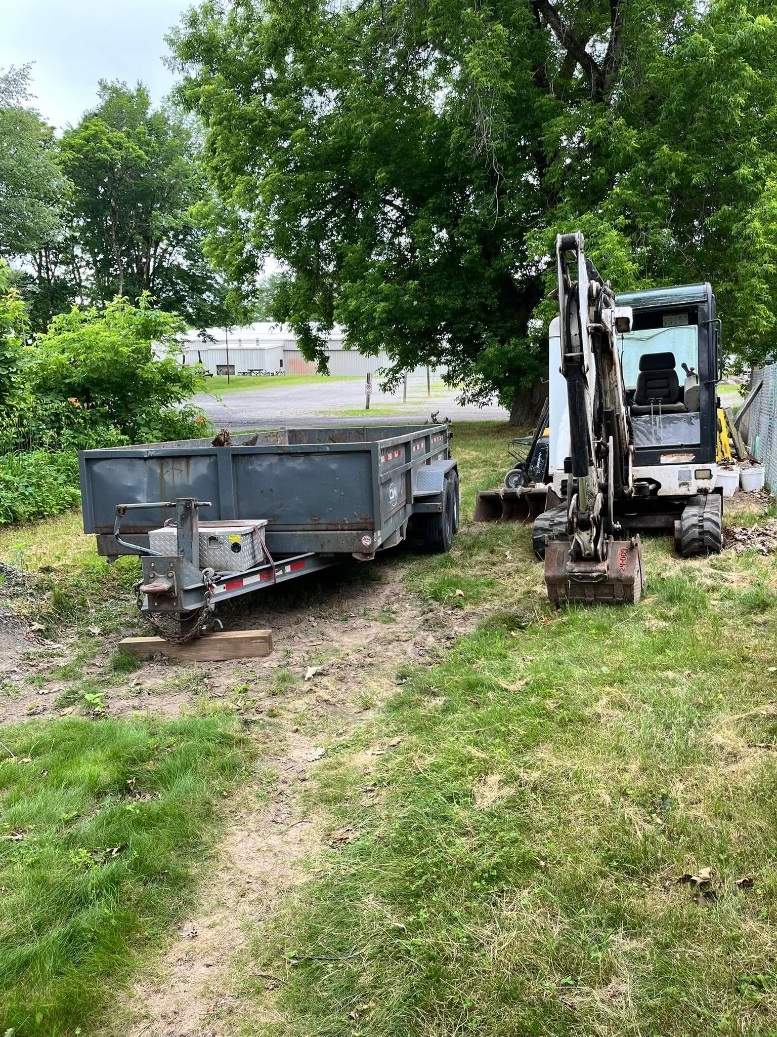 Excavator and dump trailer in a grassy yard beside trees and a driveway