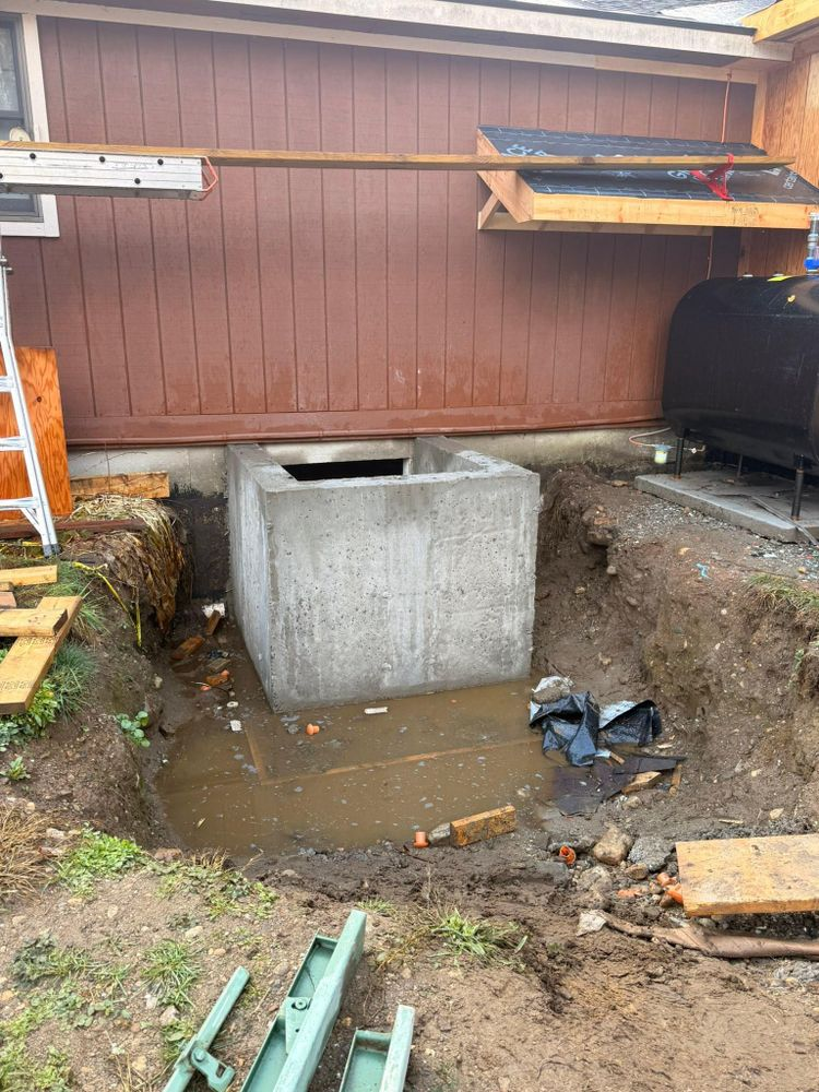 Concrete utility pit in muddy yard beside a brown building, with tools and wood planks nearby.