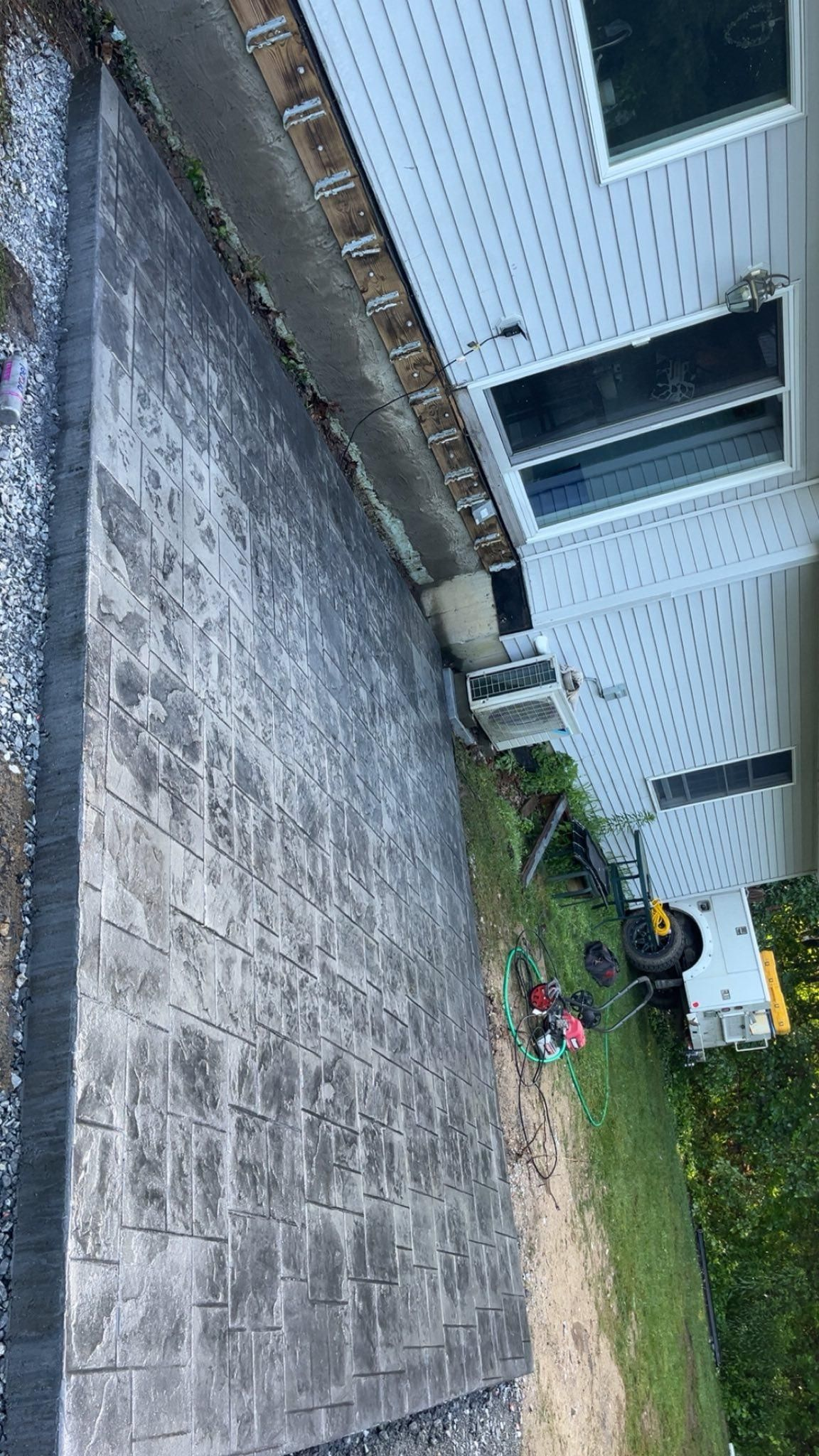Tilted view of a house side yard with concrete patio, gravel path, white siding, and bushes near a window