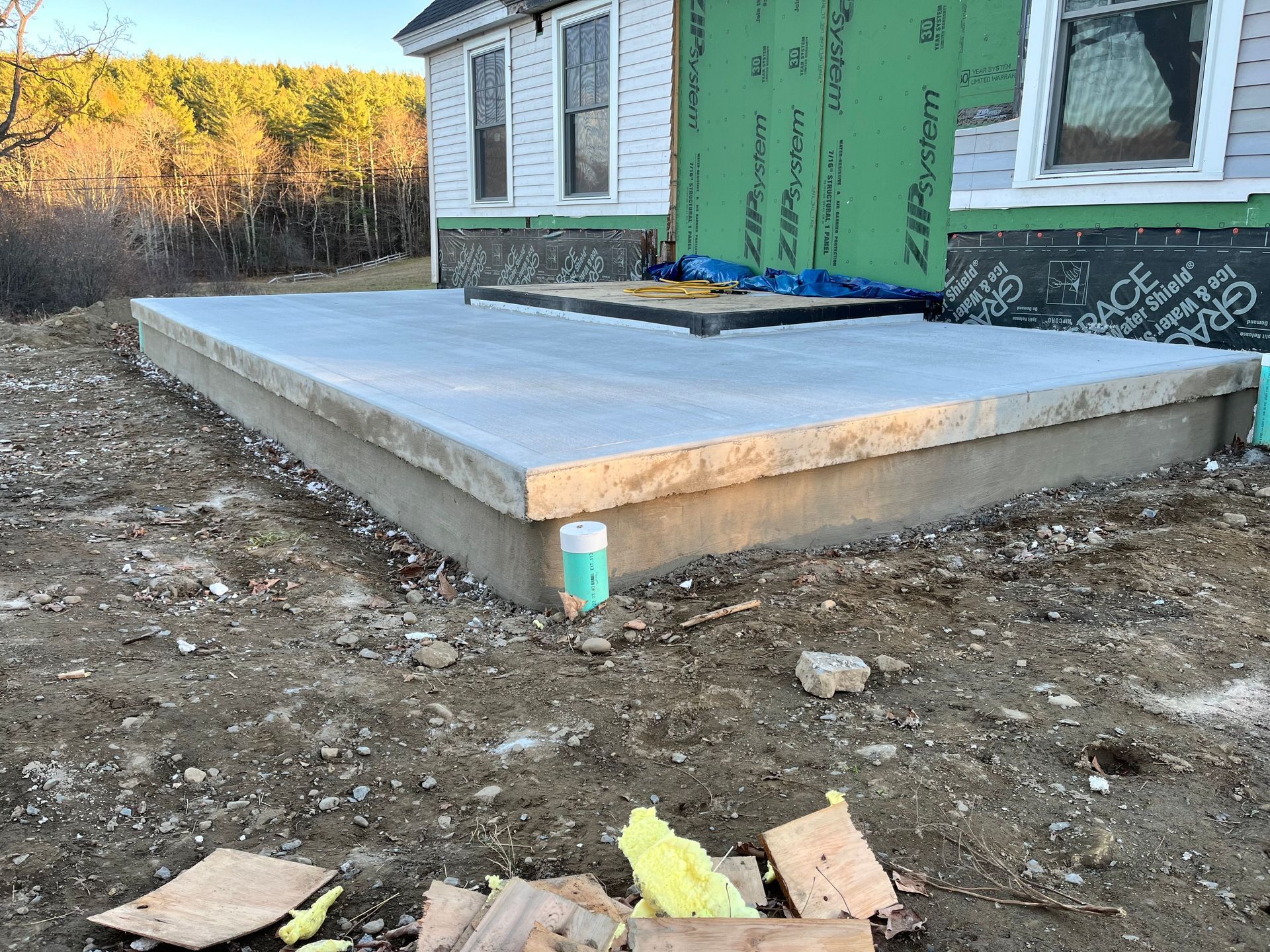 Concrete slab foundation beside a house under construction, with green house wrap and dirt foreground.