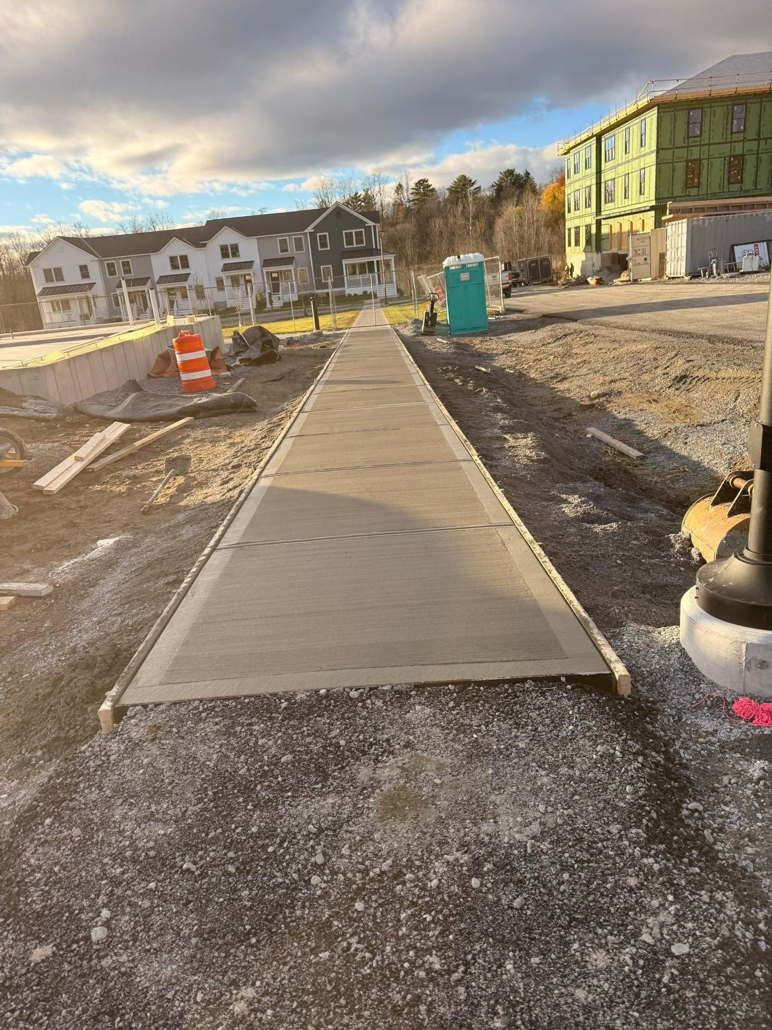 Concrete sidewalk under construction in a new housing area, with orange barrels and a green dumpster.