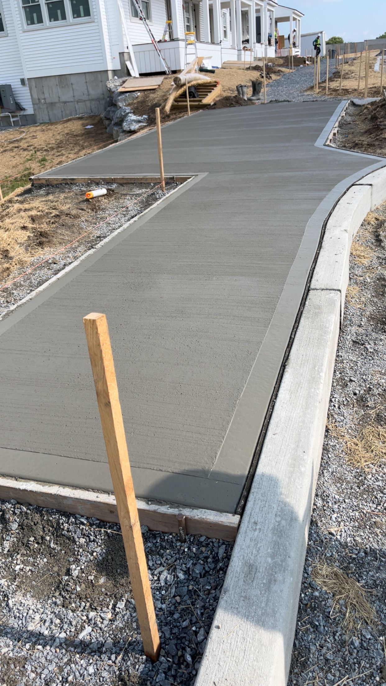 Fresh concrete sidewalk curving past houses under construction, with stakes marking the path and a curb on the right