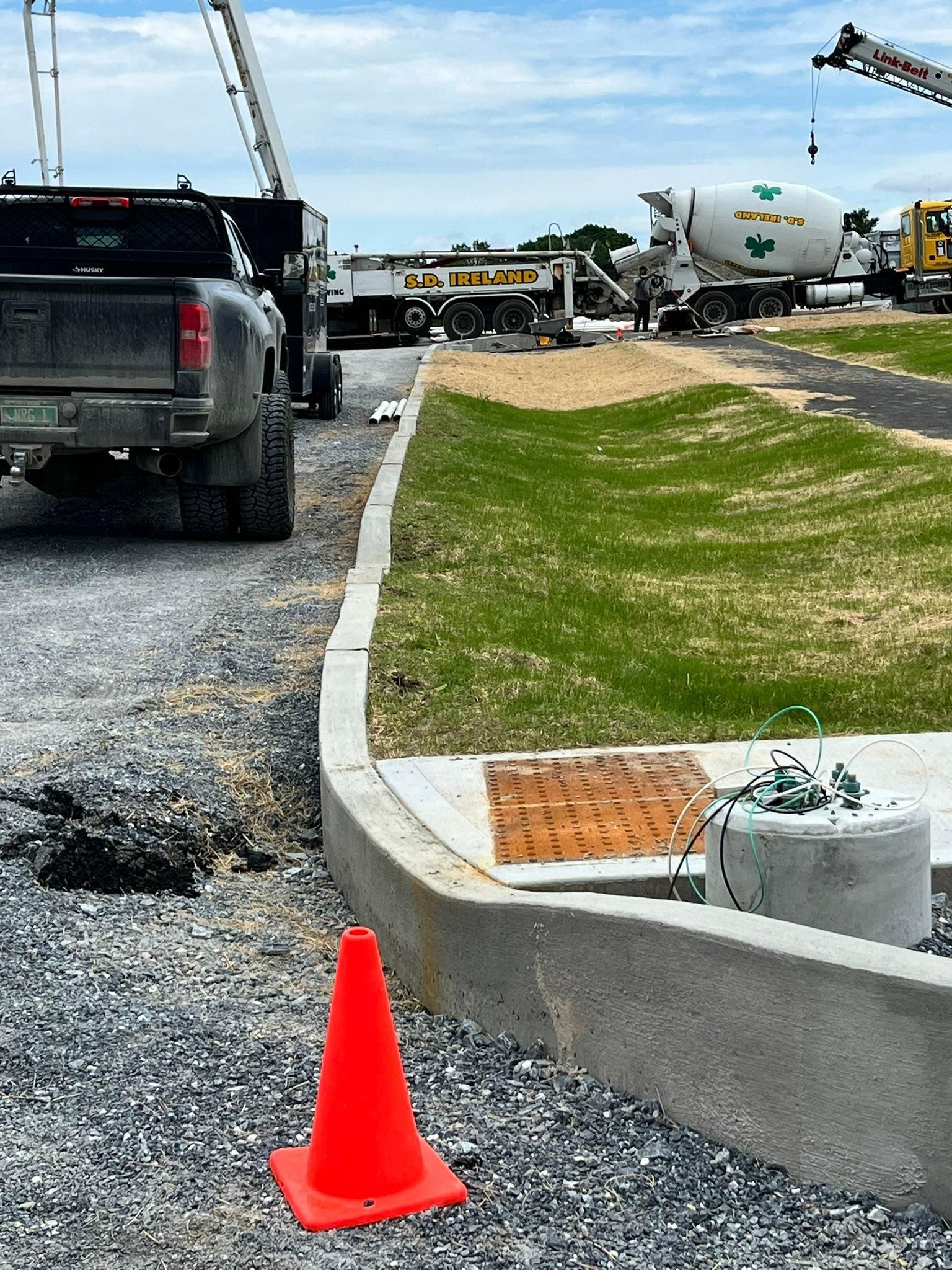 Construction site with truck, cement mixer, orange cone, gravel road, and curb along a grassy edge