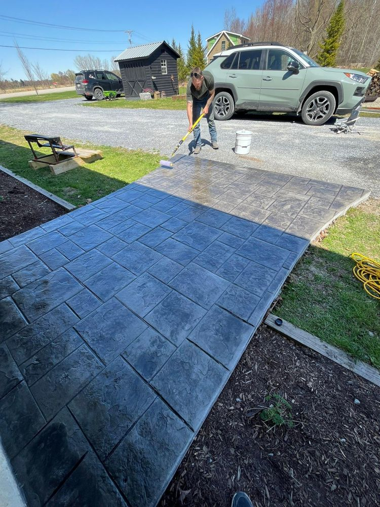 Worker staining a concrete walkway beside a driveway with a parked SUV nearby