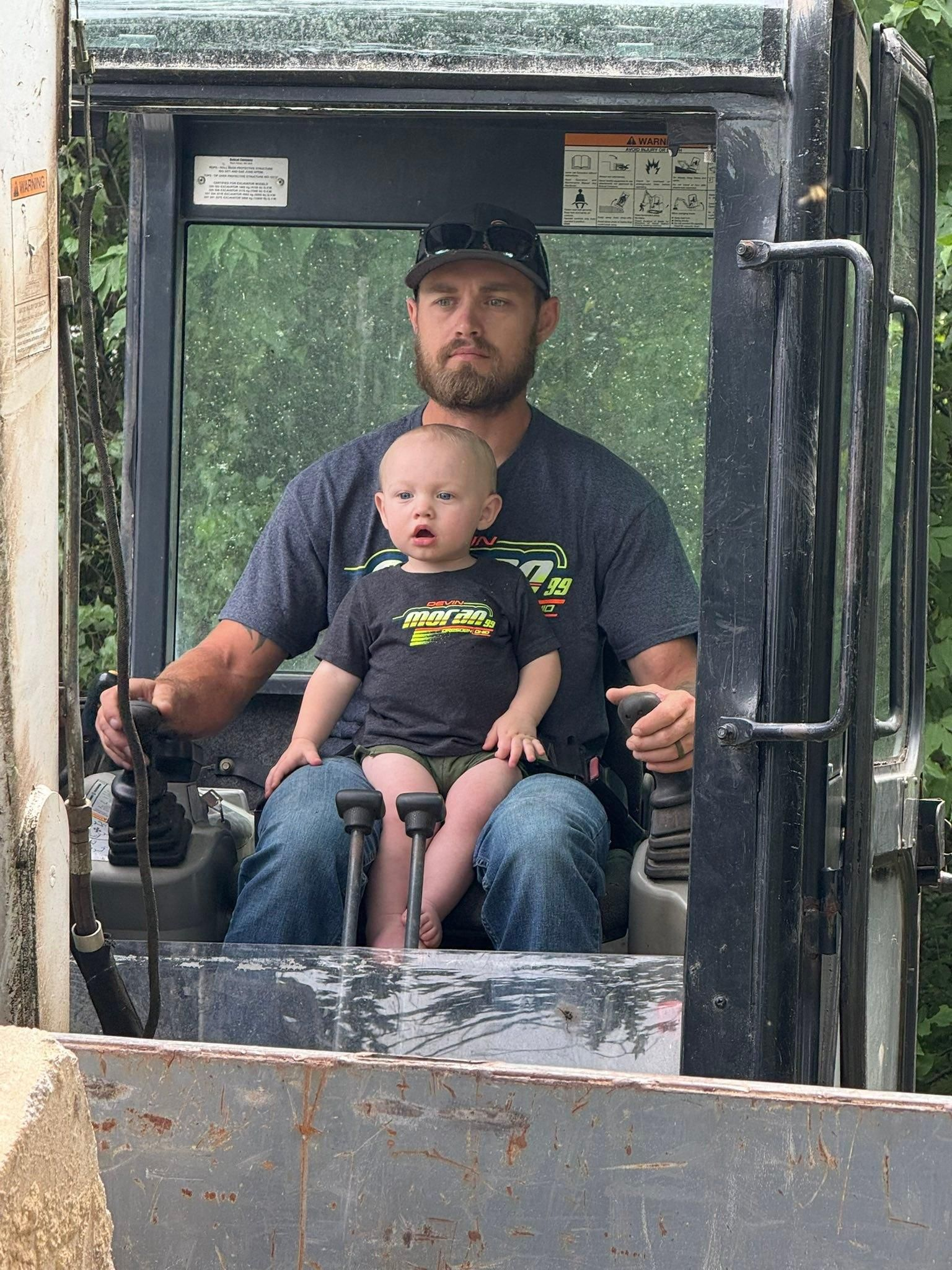 Two people seated in a bulldozer cab, one holding the controls, both facing forward.