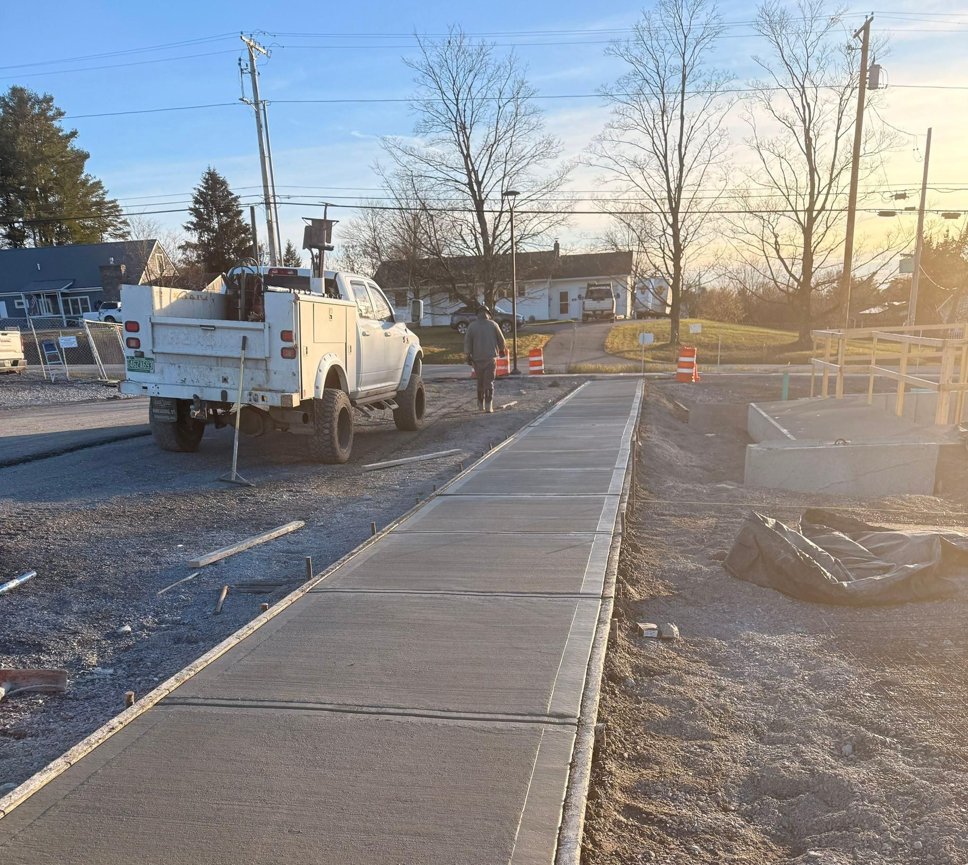 Sidewalk under construction beside a white utility truck at sunset, with cones and gravel work area