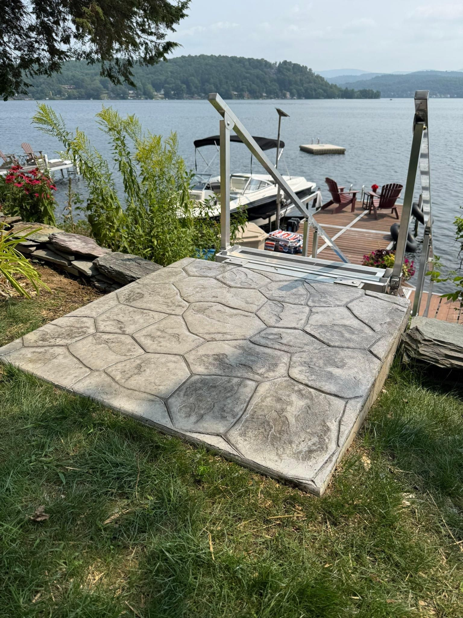 Stone path leading to a lakeside dock with chairs, boat, and green shoreline under a sunny sky