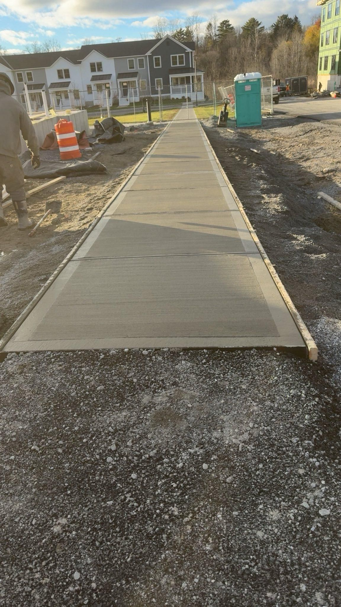 New concrete sidewalk under construction beside houses, with orange barriers and a portable toilet nearby