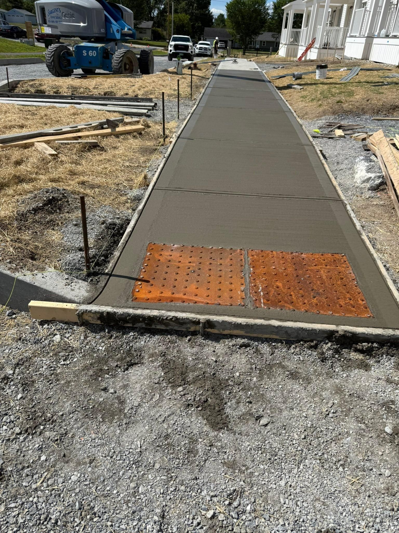 Concrete sidewalk under construction with an orange tactile warning panel at the end, gravel and equipment nearby