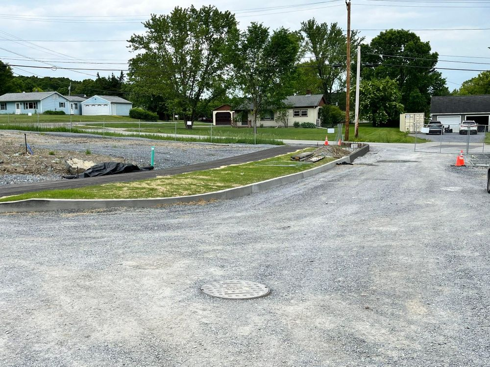 Gravel road beside a grassy lot and trees, with houses and a garage in the distance under cloudy skies.