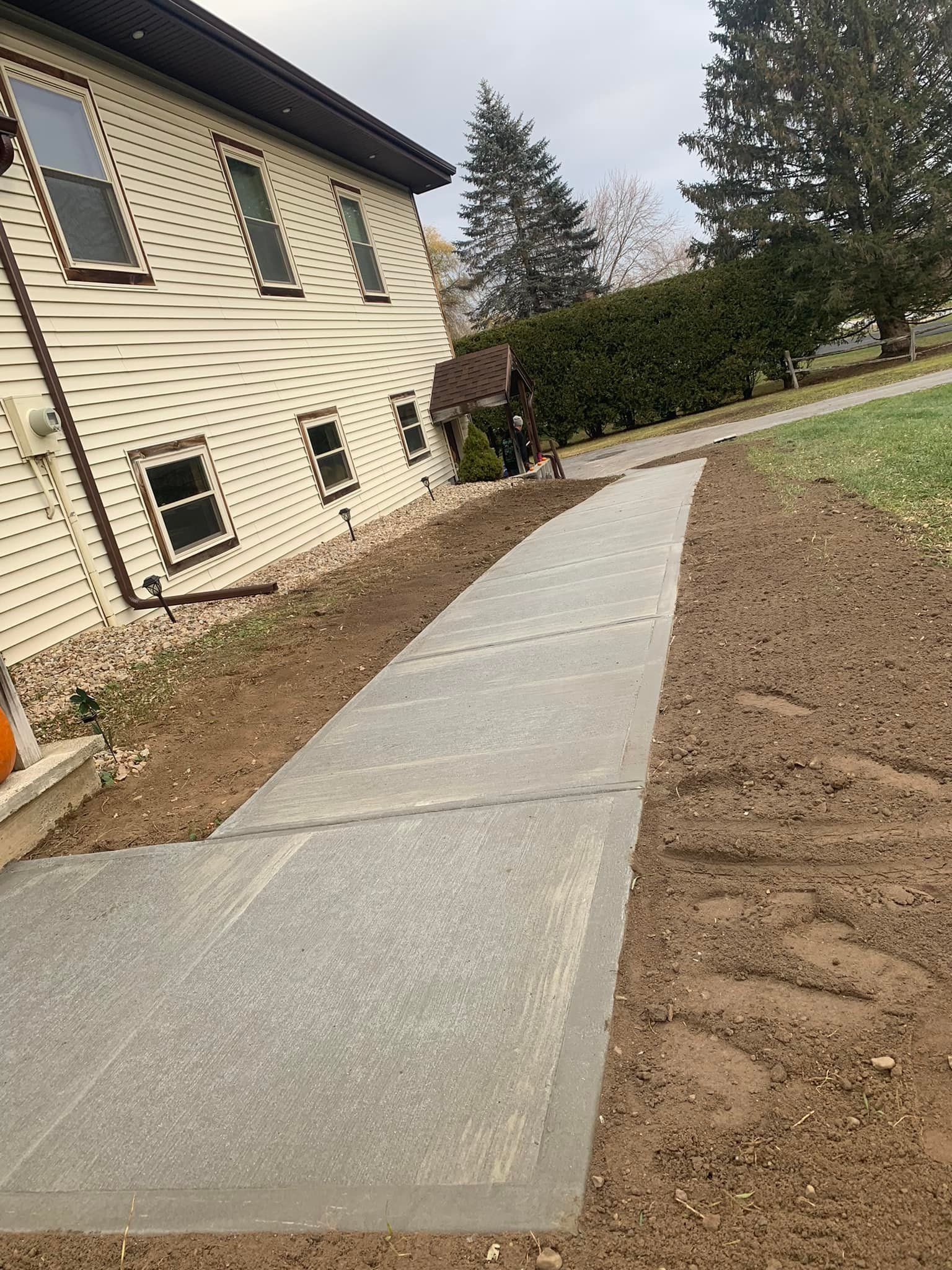 Curved concrete walkway beside a beige house, bordered by dirt and lawn, under an overcast sky
