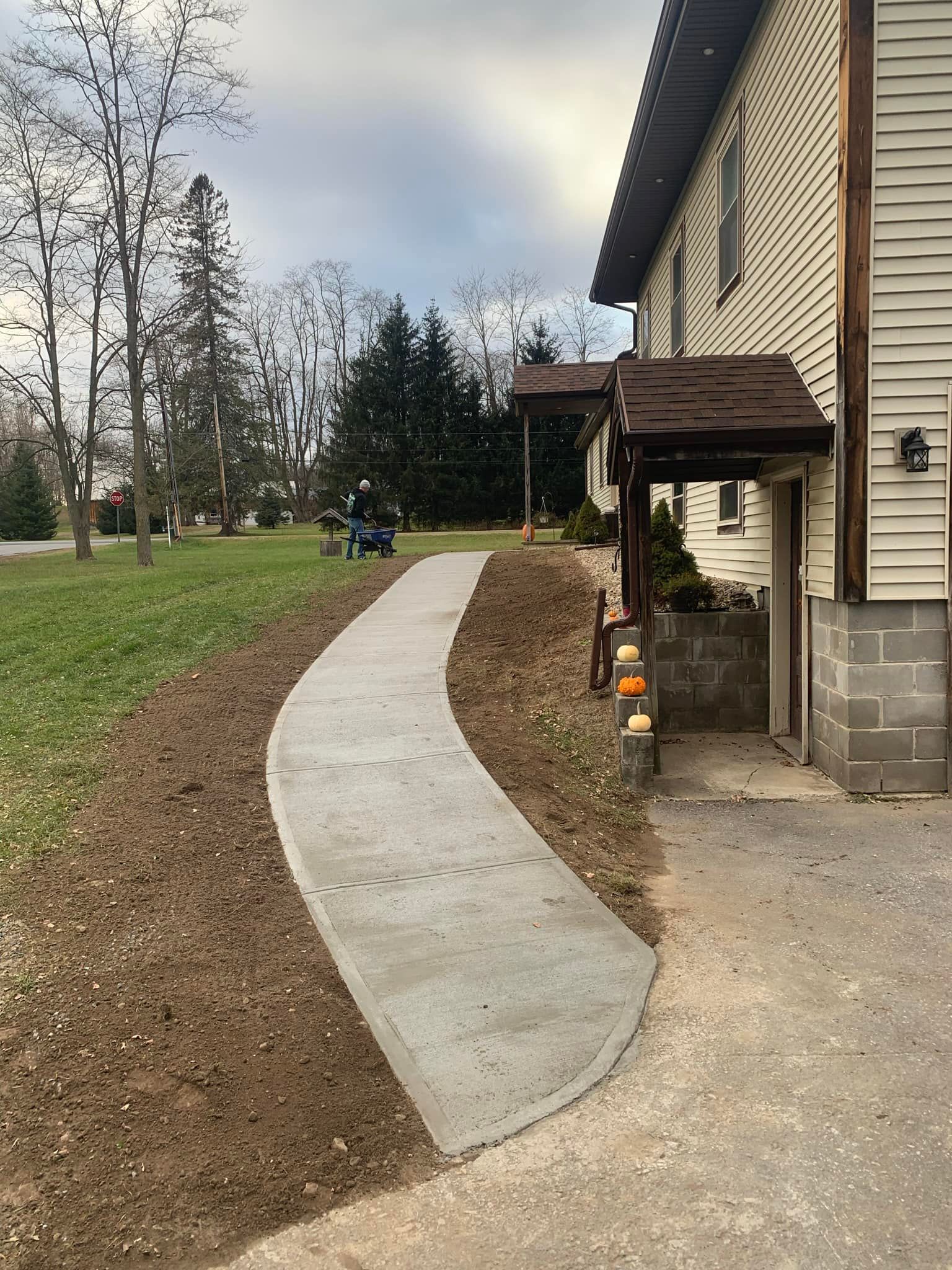 Curved concrete walkway beside a house leading through a grassy yard with trees and a cloudy sky.