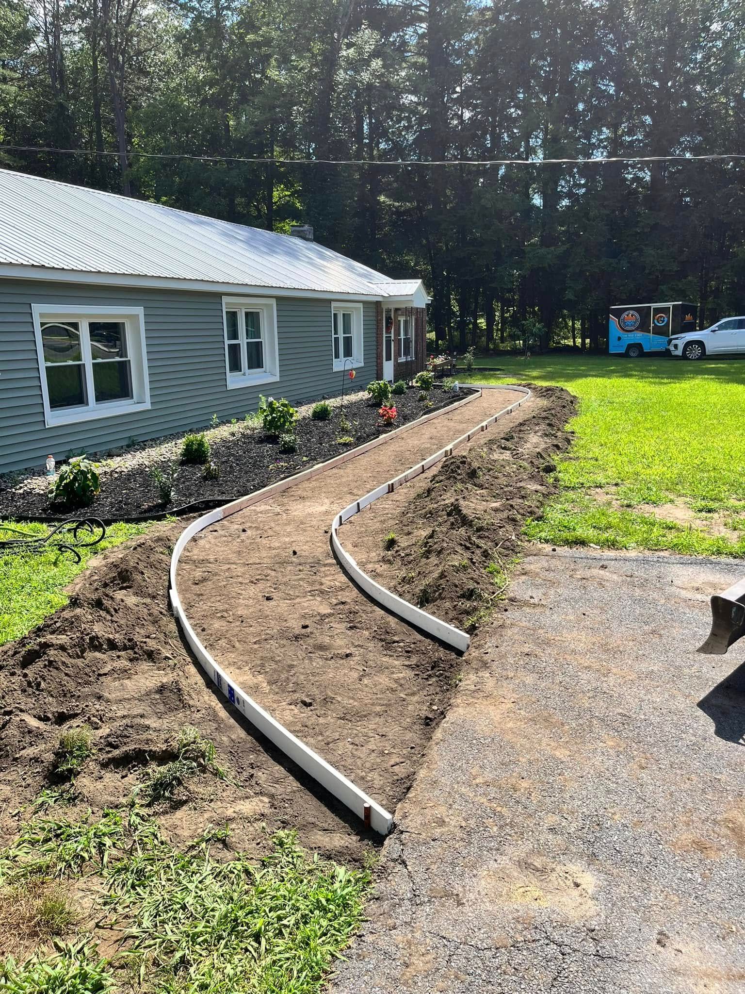 Curved garden bed under construction beside a gray house, with soil, edging, grass, and gravel driveway.
