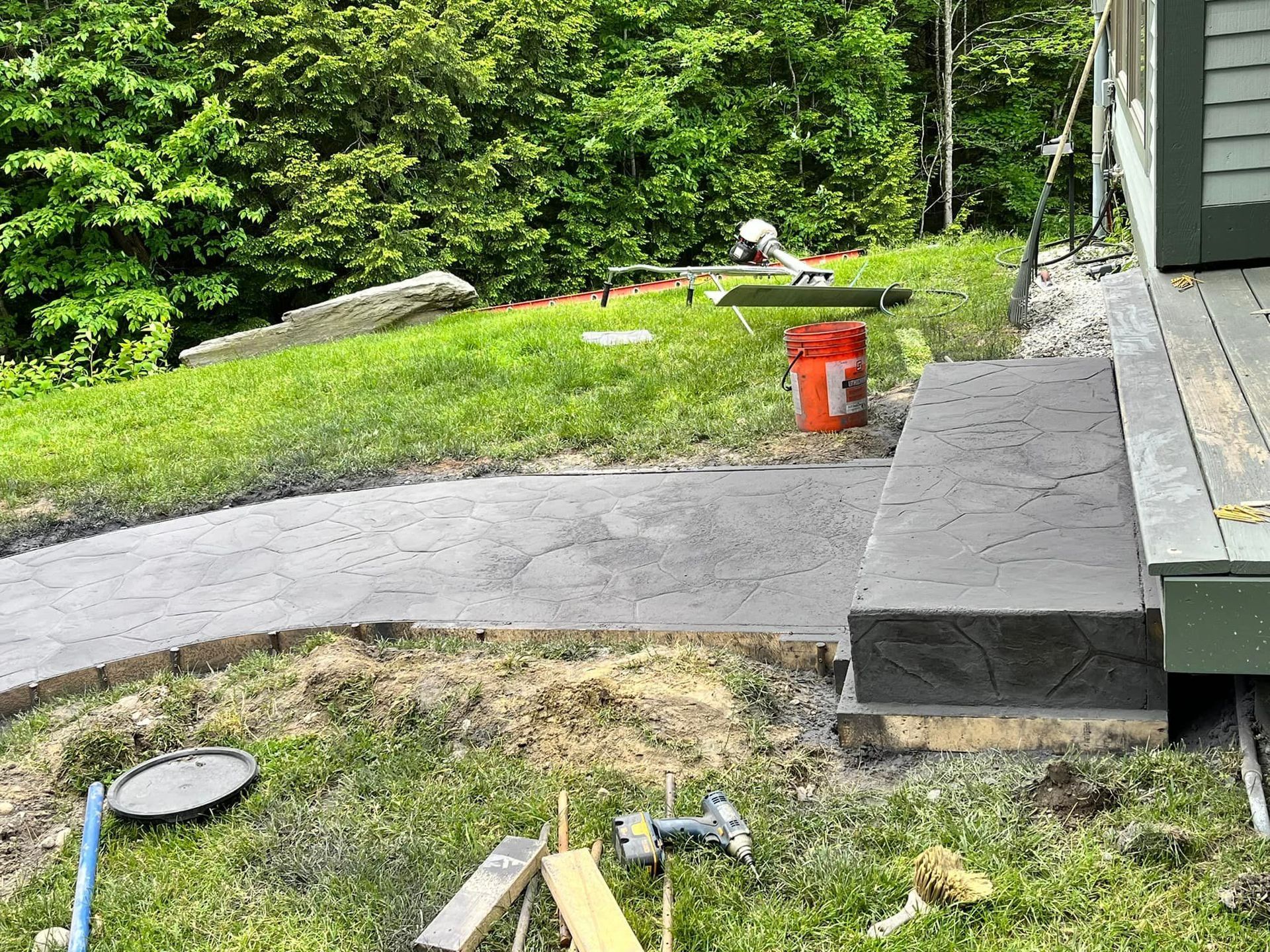 Backyard patio with curved concrete path, green lawn, and red bucket near a house exterior.