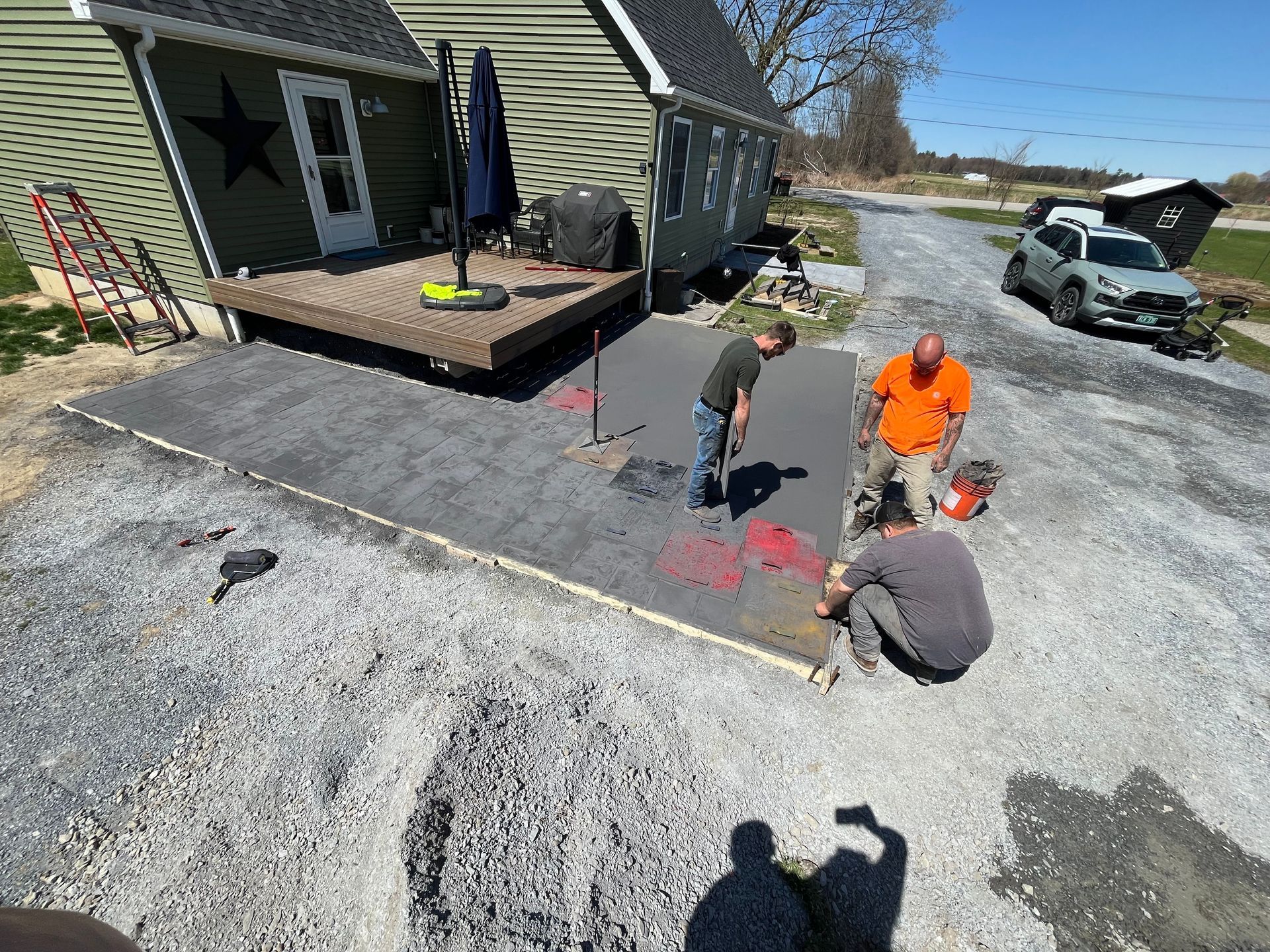 Three workers marking and leveling a gravel driveway beside a house and deck.