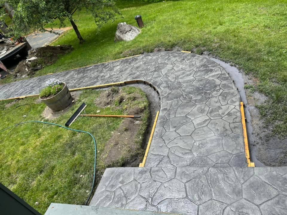Curved concrete walkway under construction with fresh stamped surface, edging strips, and tools on grass.