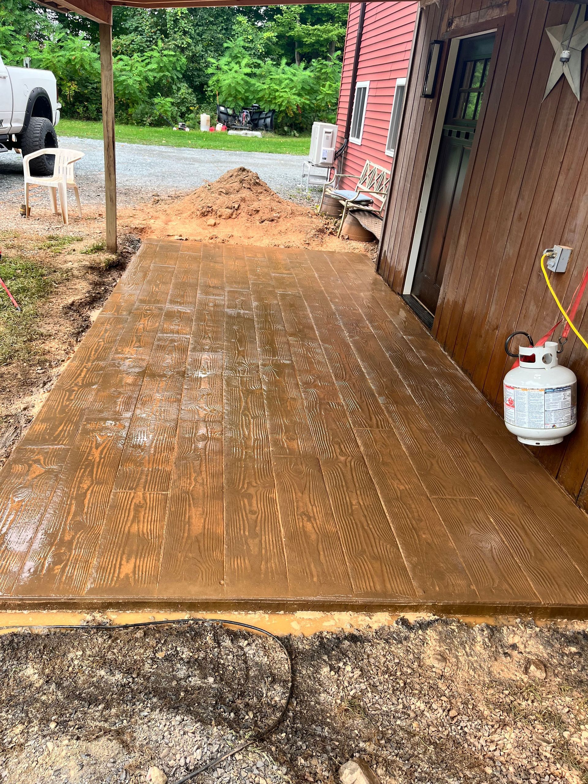 Freshly poured concrete walkway beside a brick house, with dirt pile at the end and a propane tank nearby