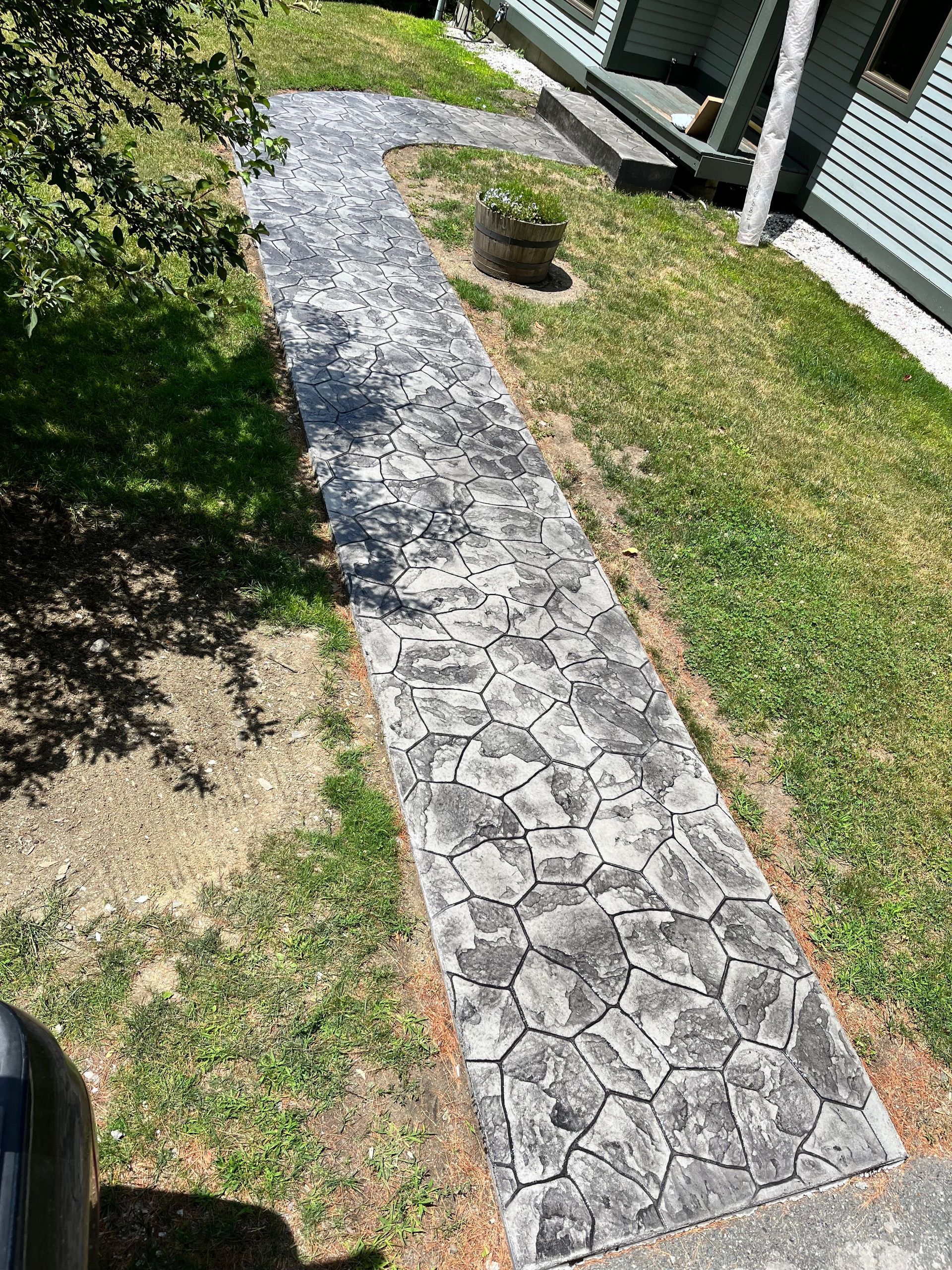 Narrow stone walkway through grass beside a white house, bordered by hedges and a small garden bed.