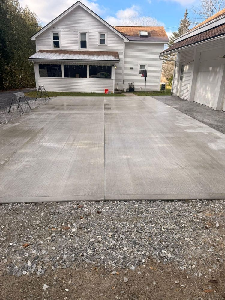 Concrete driveway in front of a white house with a garage and side building, with a gravel edge in the foreground