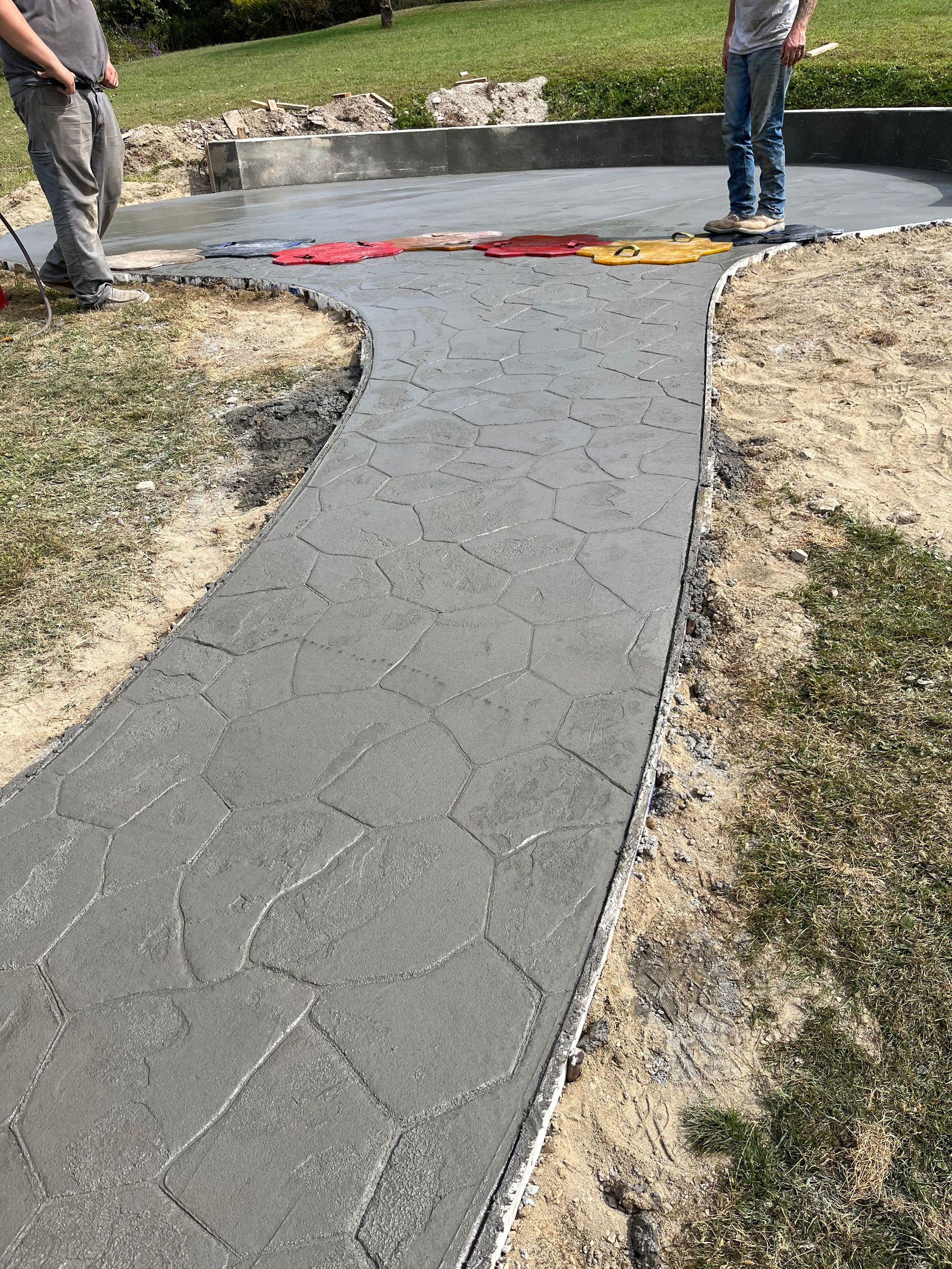 Freshly paved sidewalk curving through a grassy area, with workers standing nearby and caution tape across the top.
