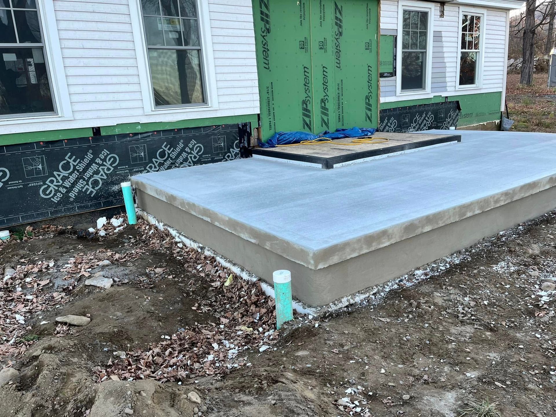 Concrete house foundation slab under construction beside a white house, with green utility pipes and exposed dirt.