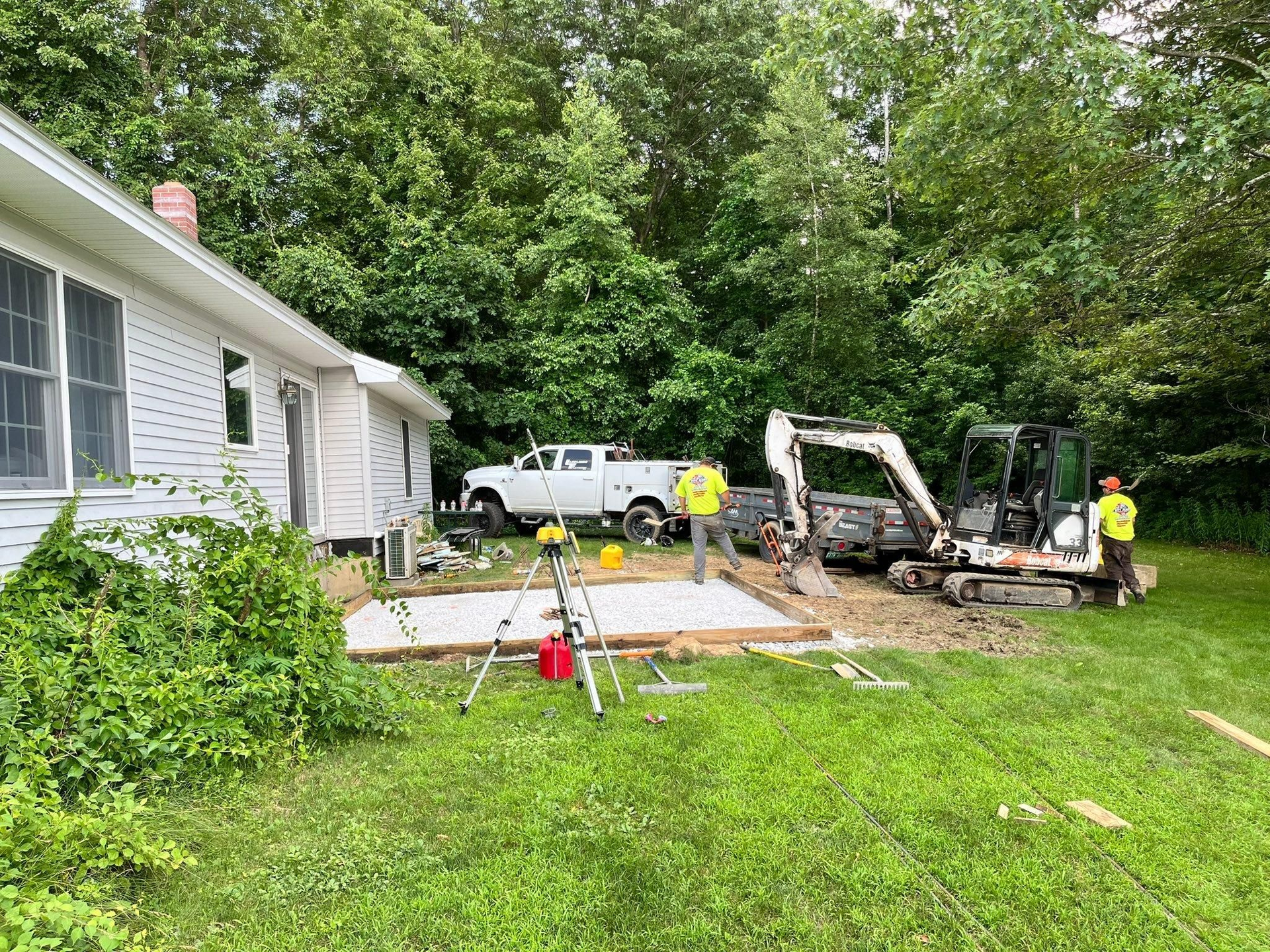 Workers with a backhoe and wheelbarrow repairing a patio beside a house in a wooded yard.