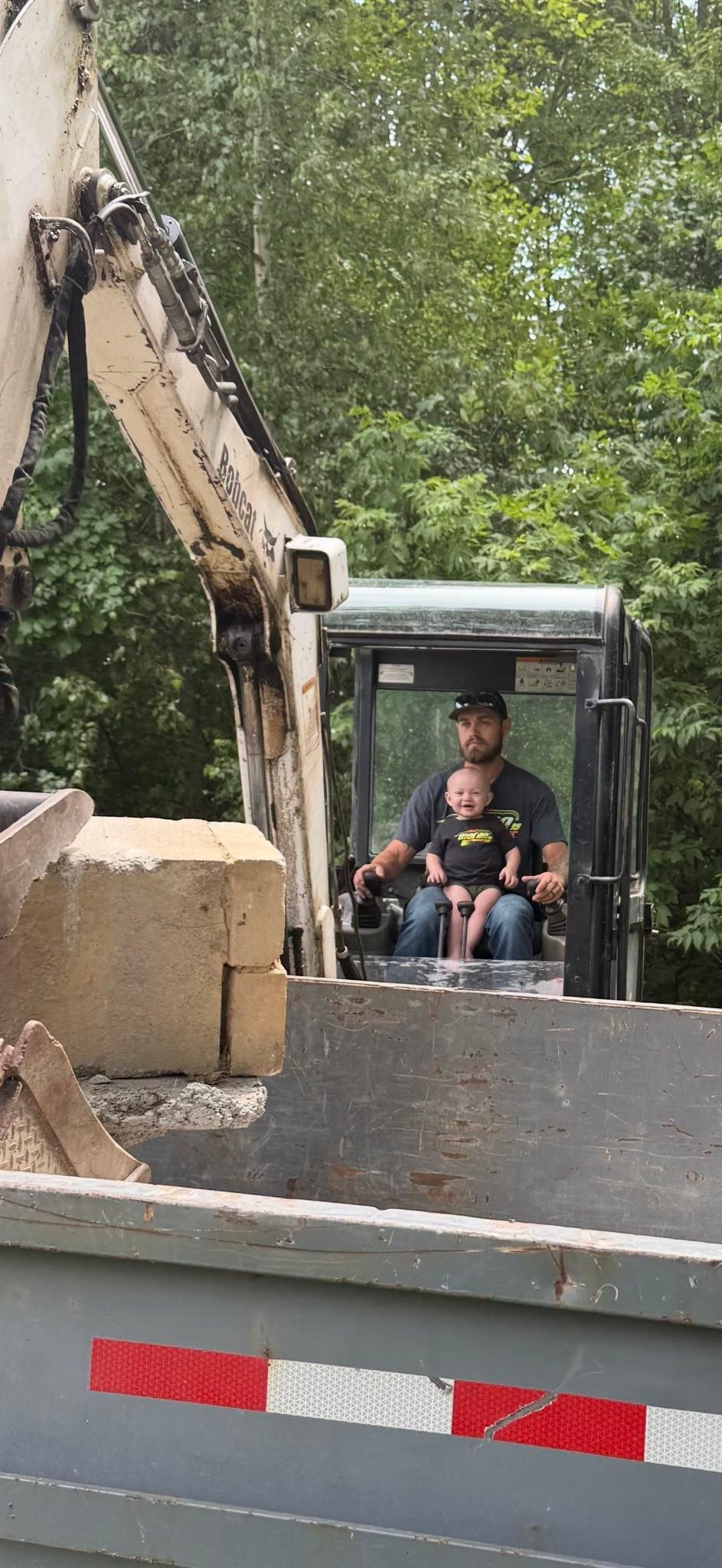 Excavator digging beside a road, with a worker seated in the cab near a wooded area.