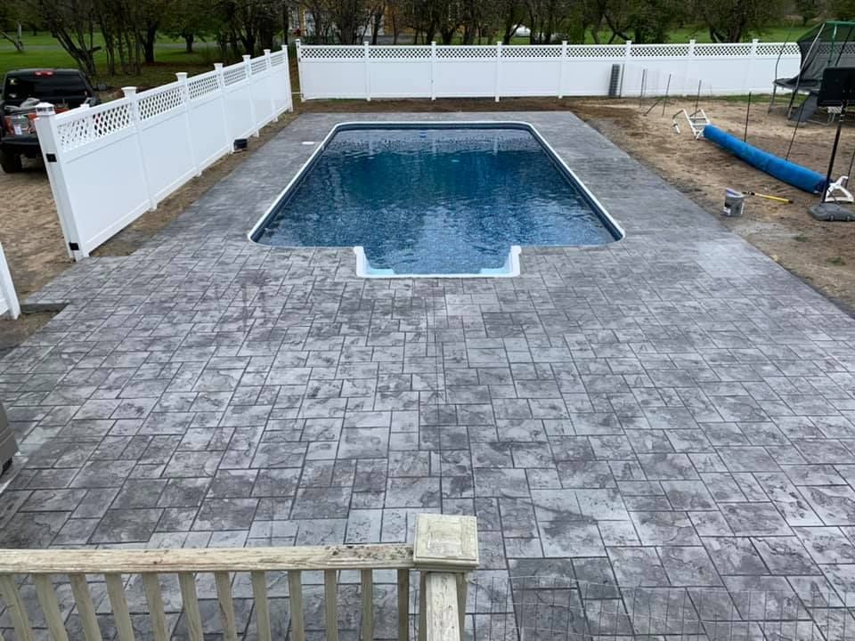 Backyard with an empty rectangular swimming pool and a large stone patio, bordered by white fencing.