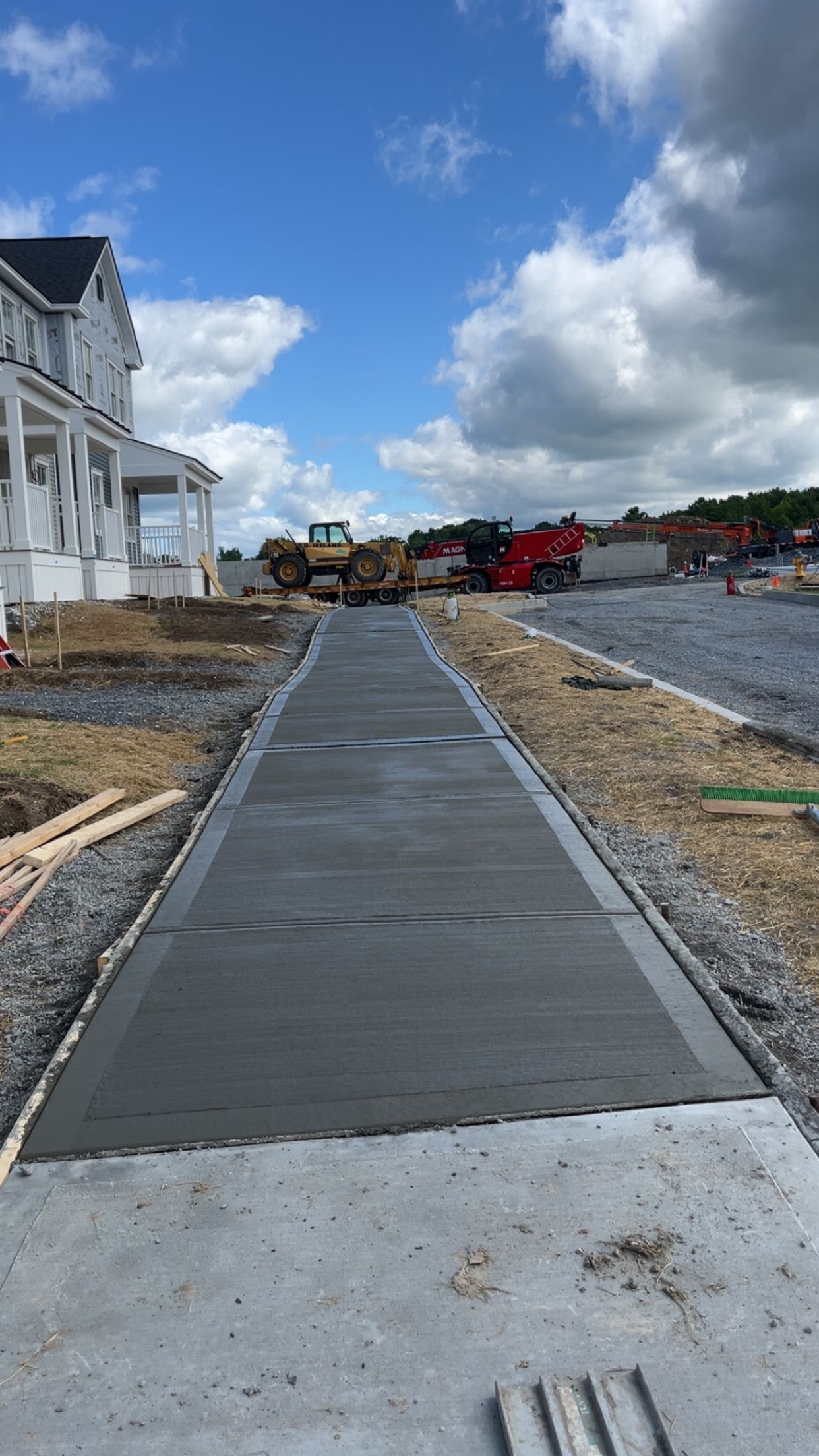 Concrete walkway under construction beside houses and gravel lot, with heavy equipment in the distance.