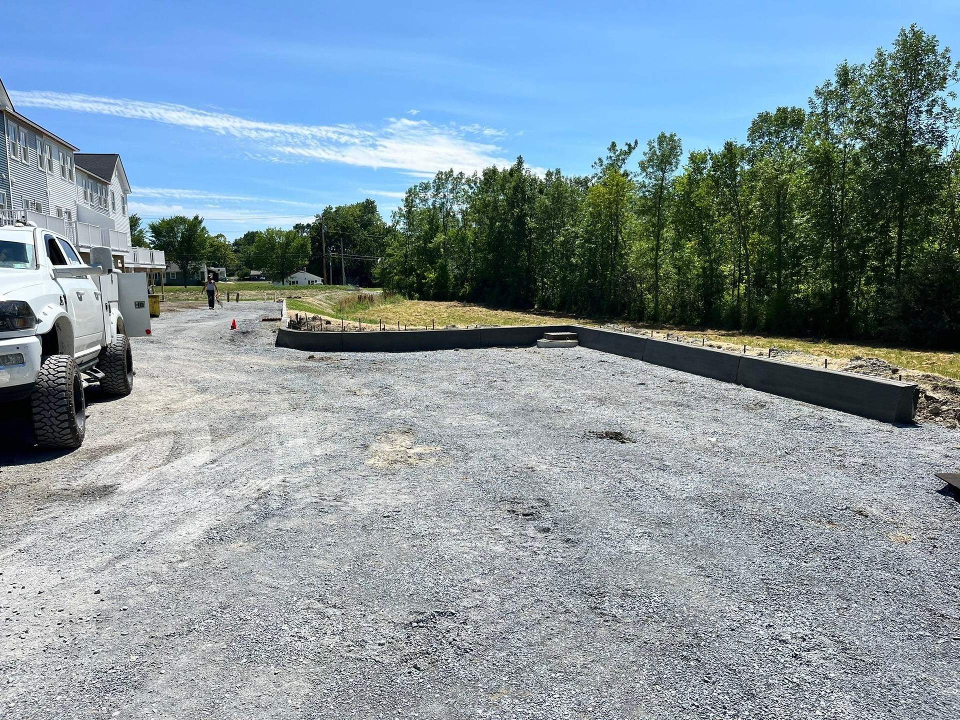 Gravel lot with concrete curbs, parked trucks on the left, and trees lining the right side under a blue sky