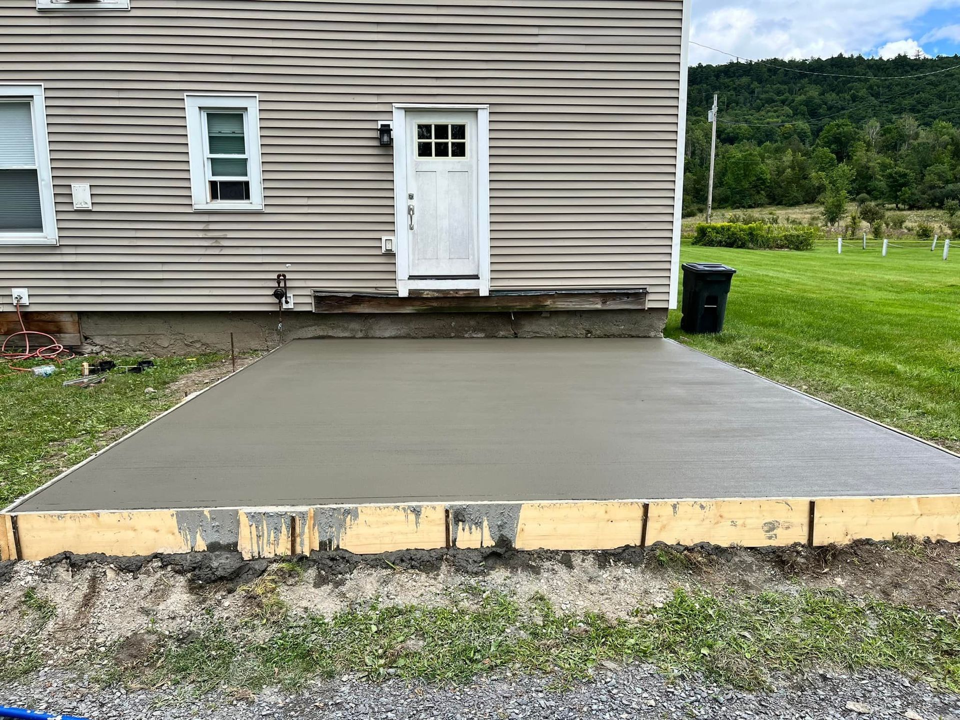 Concrete patio in front of a house with a white door and beige siding, beside a grassy yard.