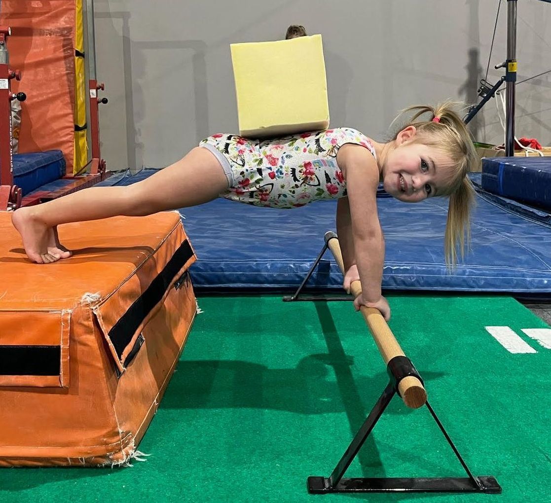 A woman is helping a young girl do a handstand on a bar in a gym.