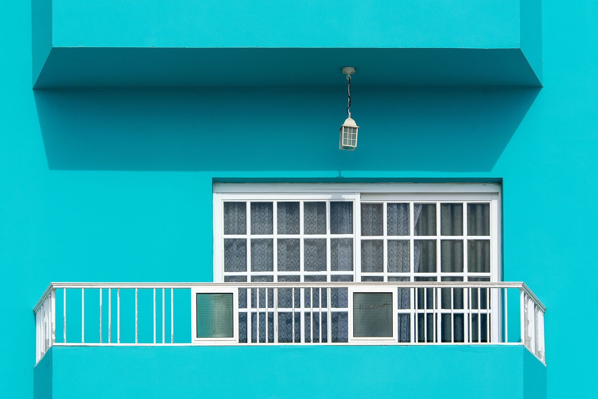 Turquoise building facade with a small balcony, white railing, and a central window under an awning