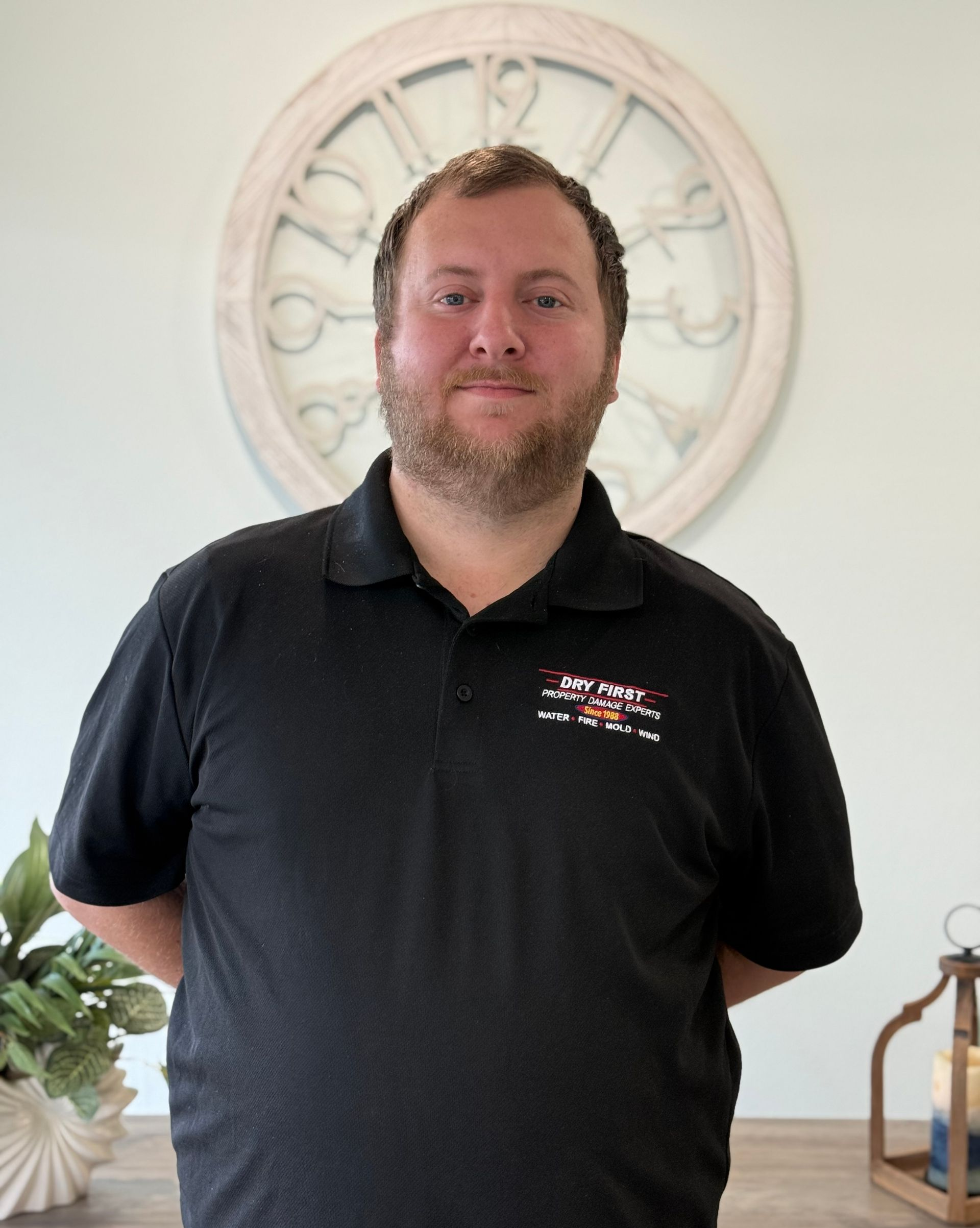 member of dryfirst restoration company with a beard stands in front of a wall clock, wearing a black polo shirt with a logo.