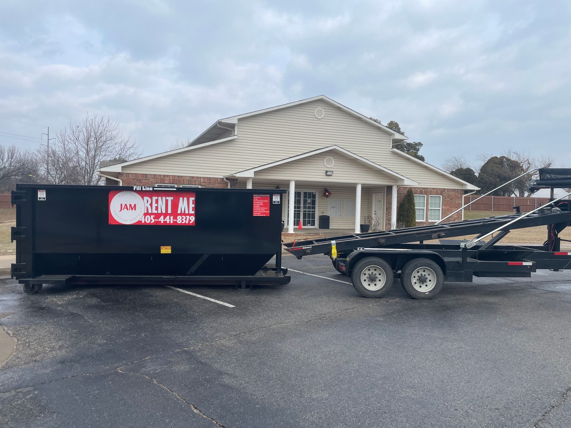 A dumpster is being towed by a trailer in front of a house.